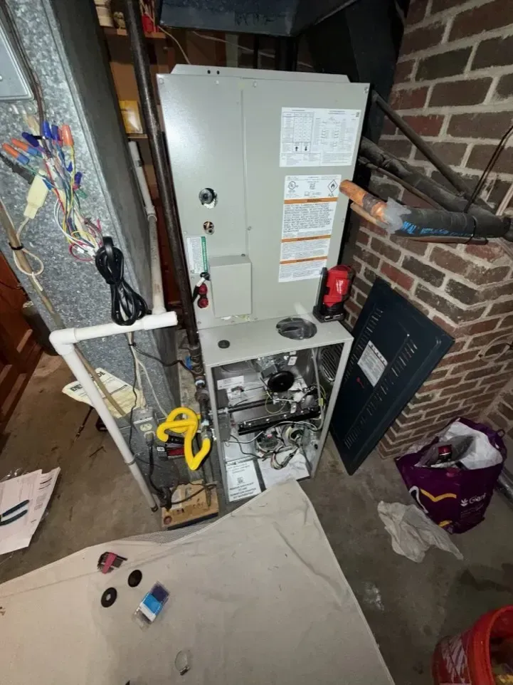 Utility closet with open furnace panel, pipes, and tools in a cluttered basement corner.