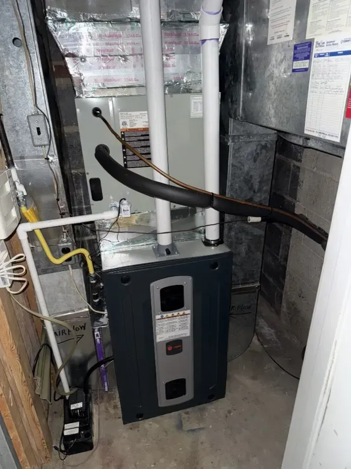 Black HVAC/furnace unit with connected pipes and ducts in a utility closet.
