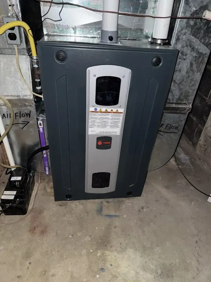 Black furnace/air handler in a utility room with pipes and concrete floor, front access panel visible