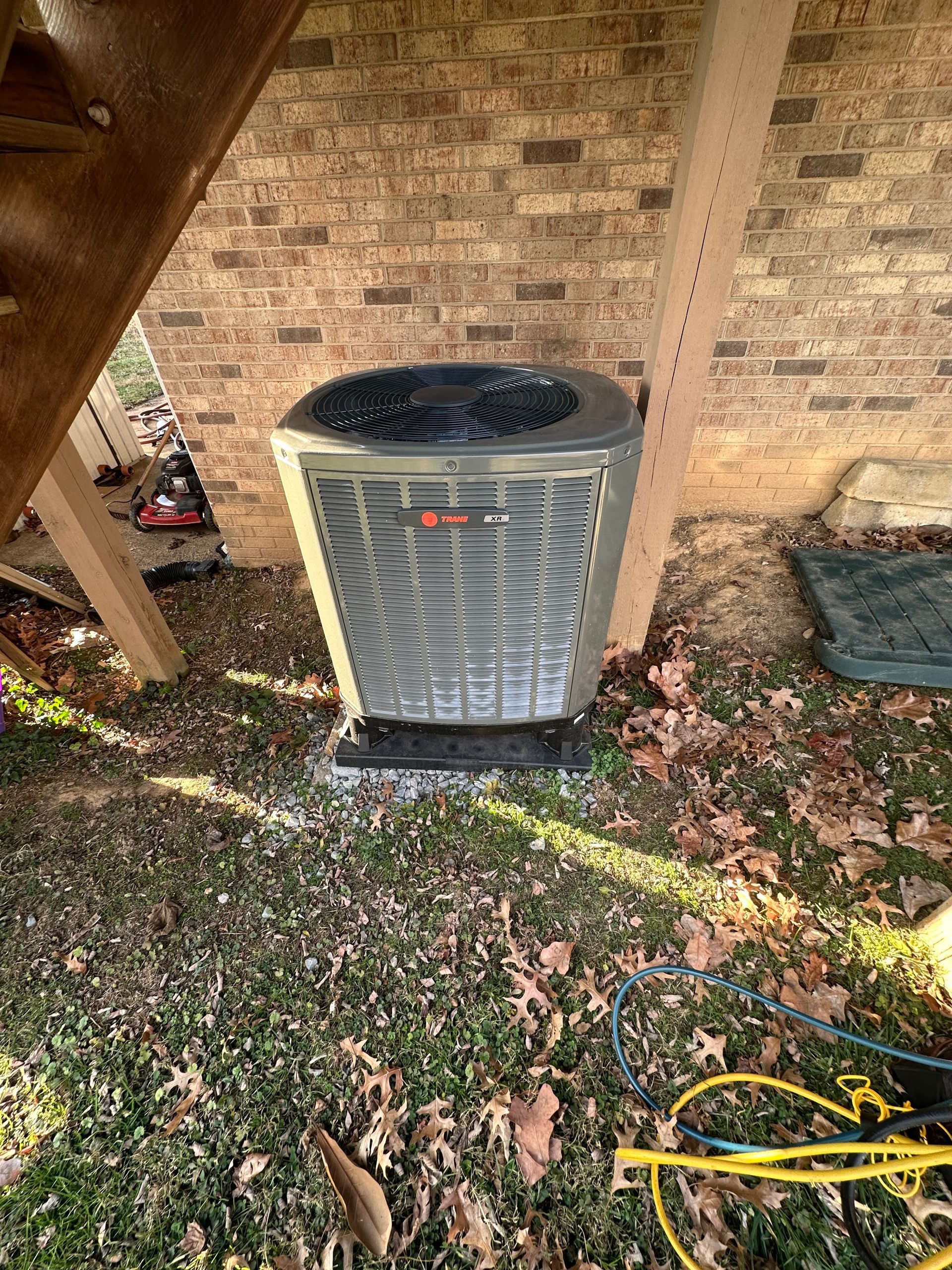 Outdoor HVAC unit beside a brick wall, with grass, leaves, and a yellow hose in the foreground.