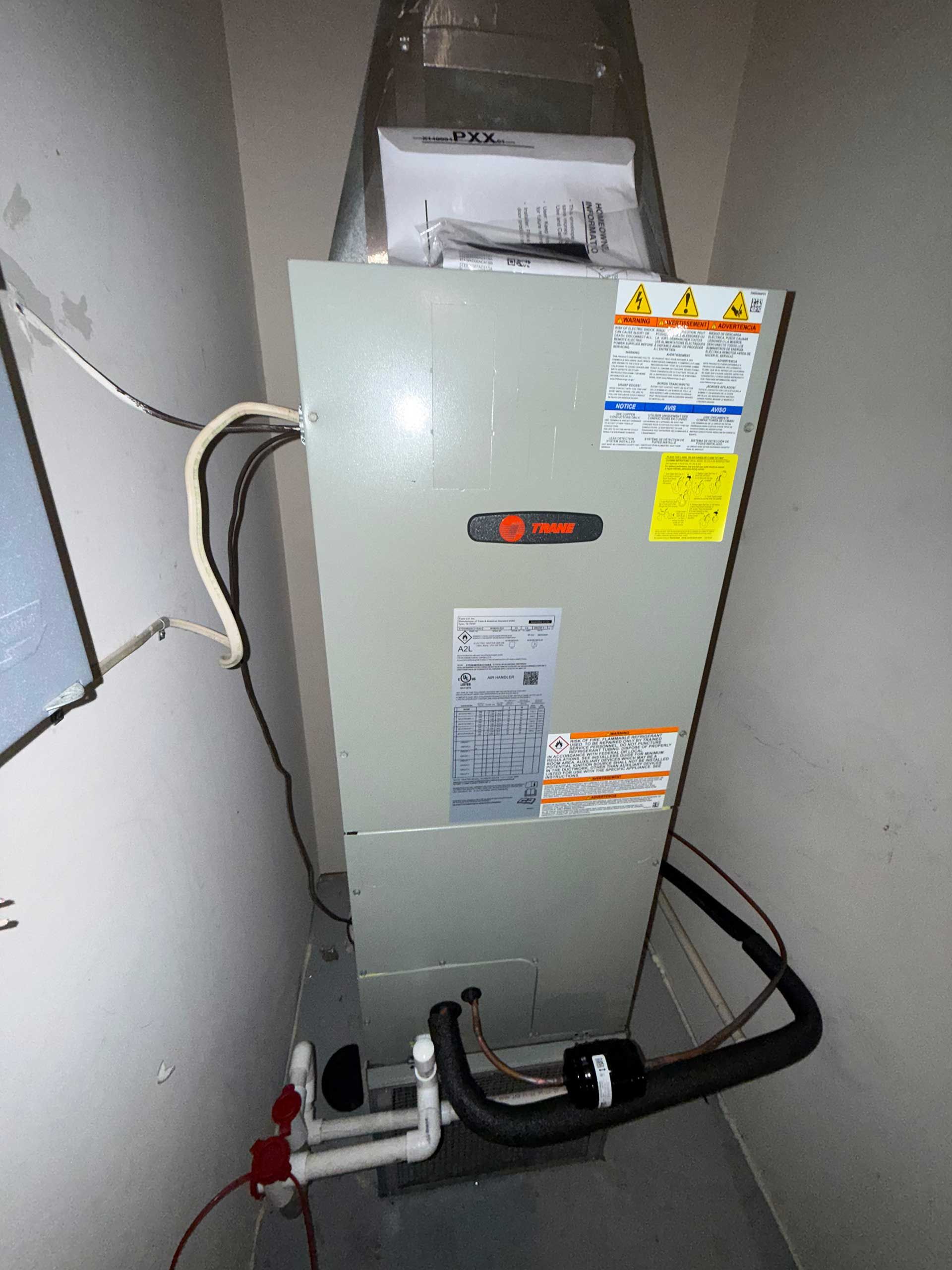 Gray furnace/air handler in a utility closet with visible pipes, wiring, and warning labels.