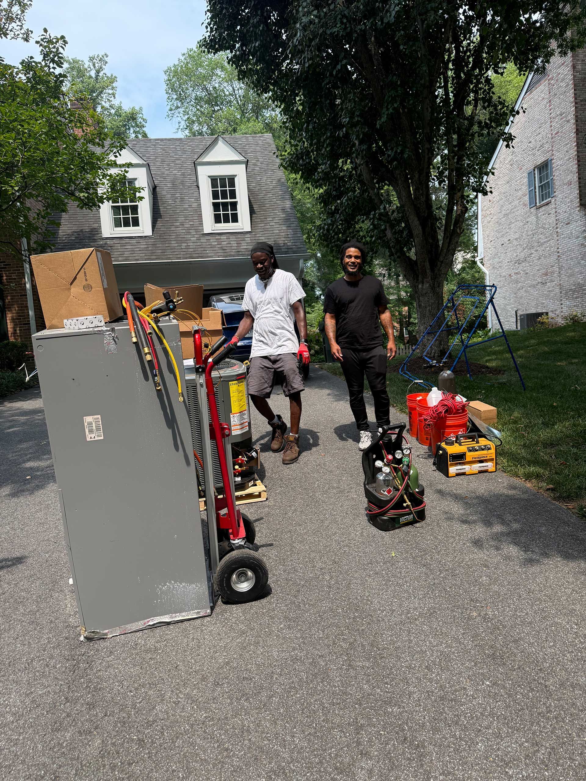 Two men standing beside lawn equipment and tools in a driveway with trees and houses in the background
