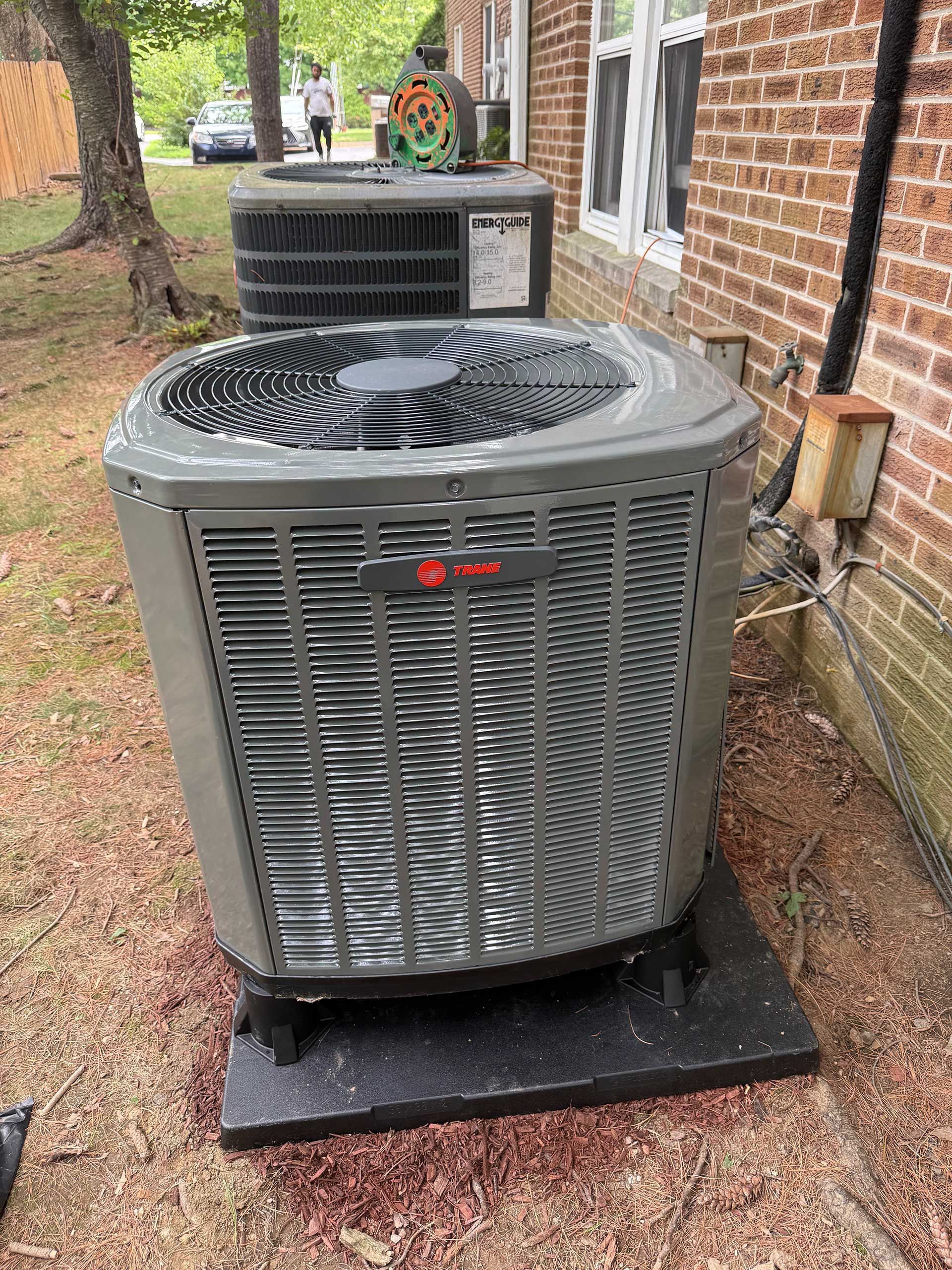 Two outdoor HVAC condenser units beside a brick house on a grassy yard.