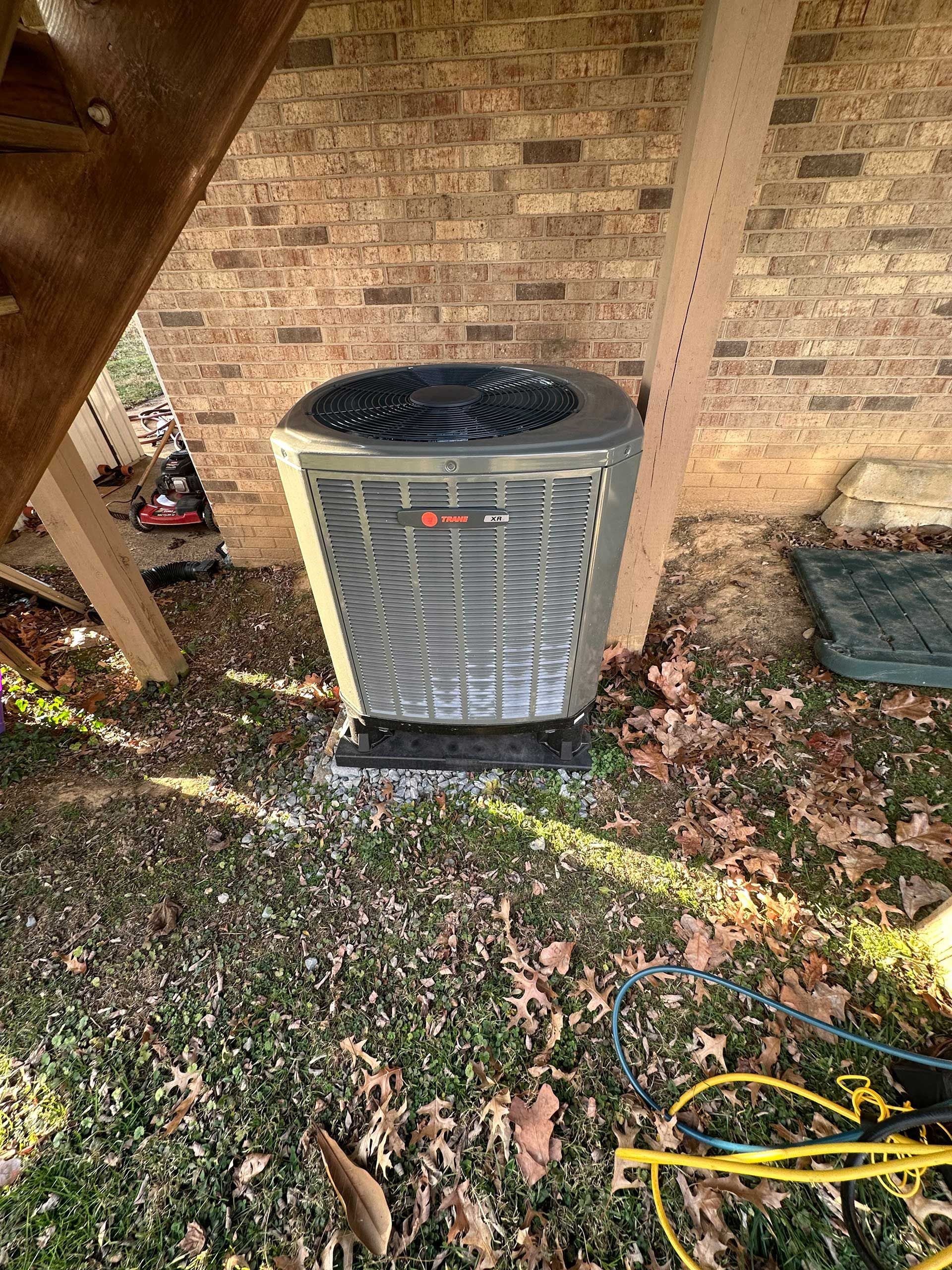 Outdoor HVAC condenser unit beside a brick wall, with leaves and a yellow hose on the ground.