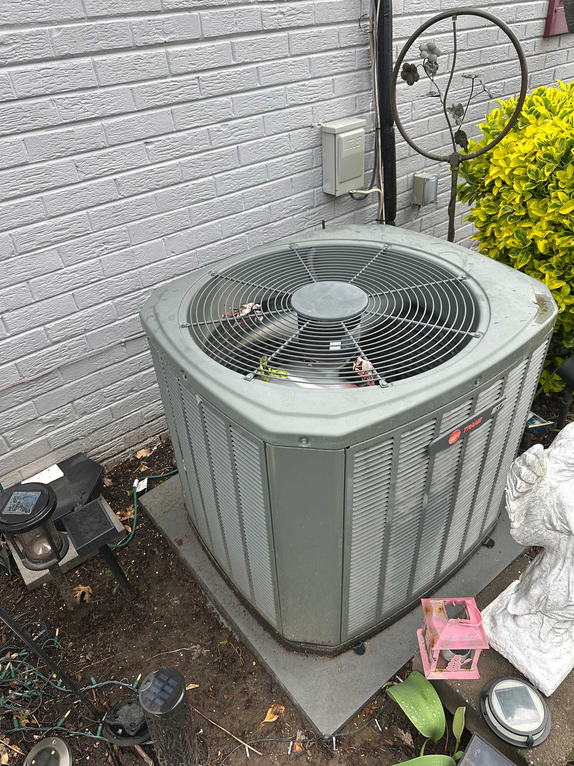 Outdoor air conditioner unit beside a brick wall, surrounded by plants and debris.