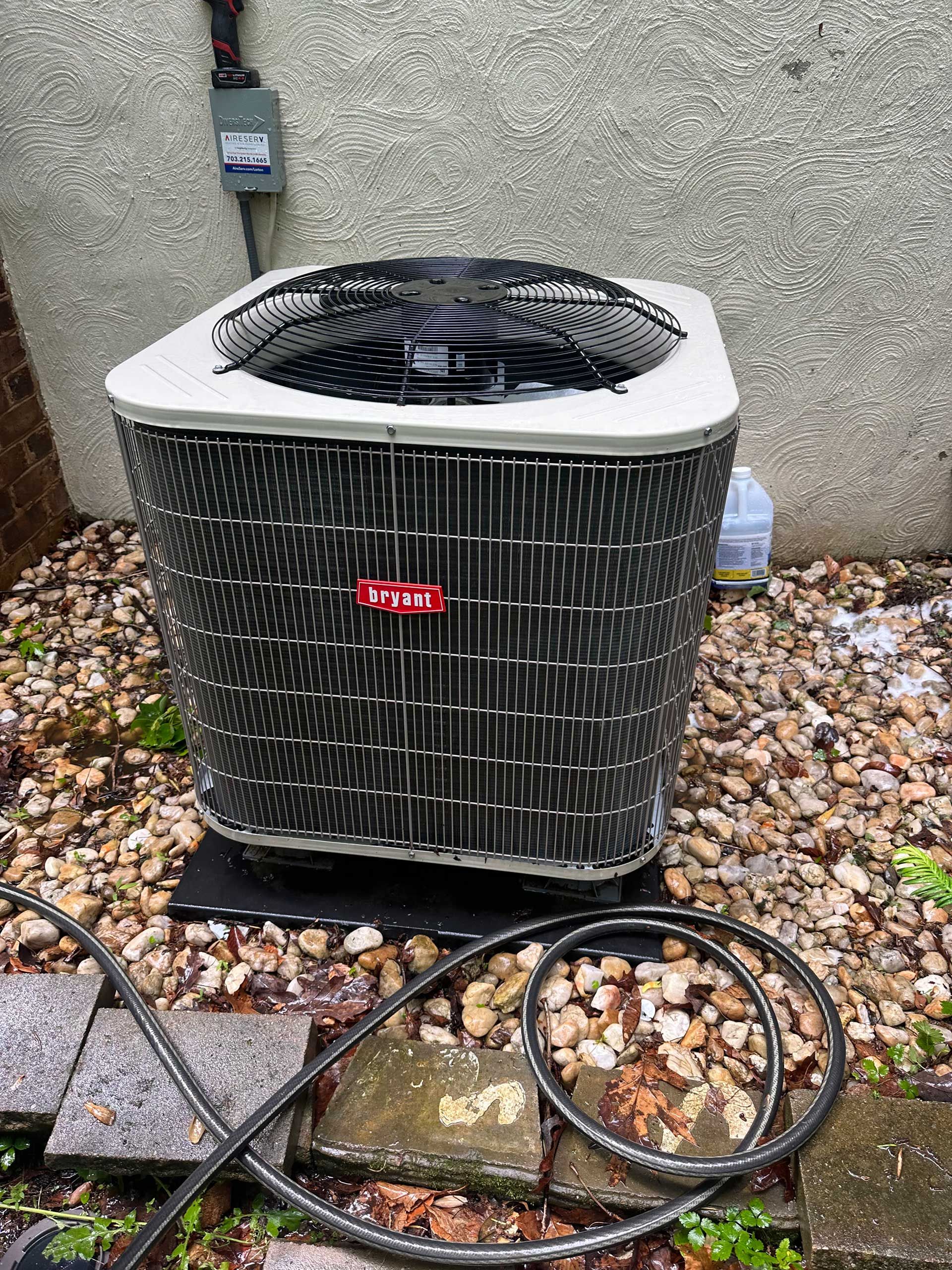 Outdoor HVAC condenser unit beside a wall, with gravel and coiled black hoses on the ground.