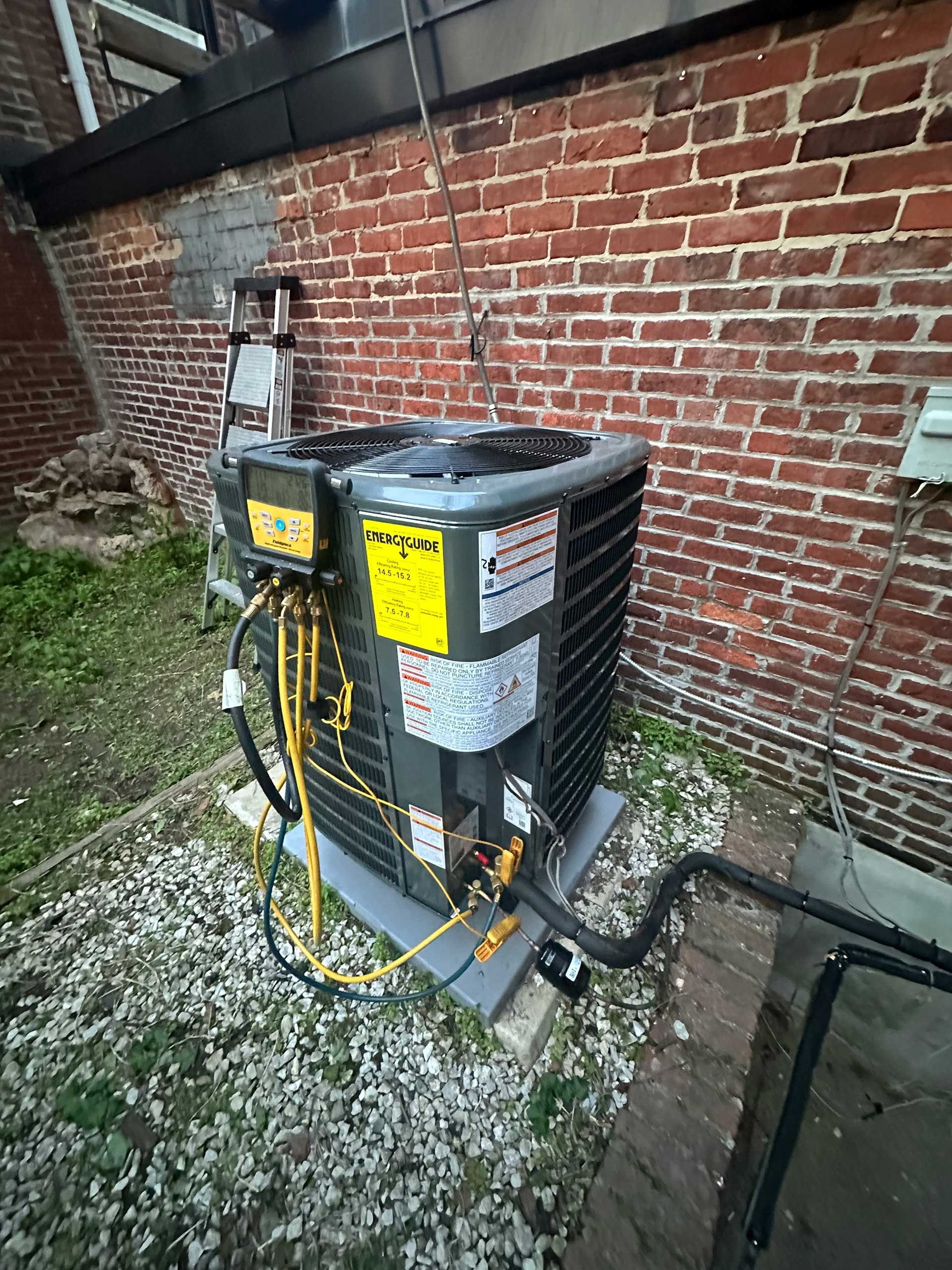 Outdoor HVAC unit beside a brick wall, with yellow warning labels and pipes on a concrete pad