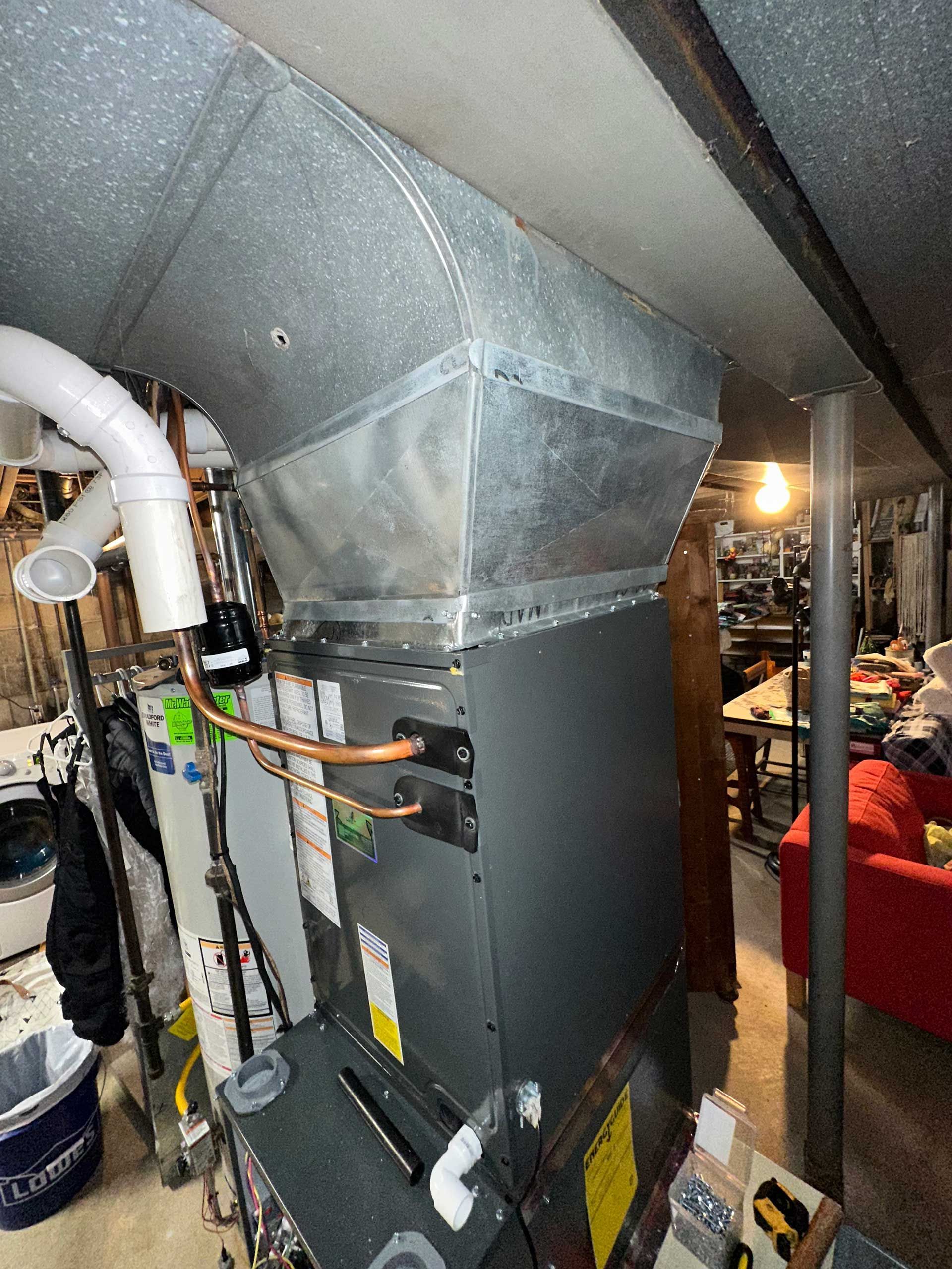 Basement HVAC ductwork and furnace next to exposed pipes and a cluttered room.
