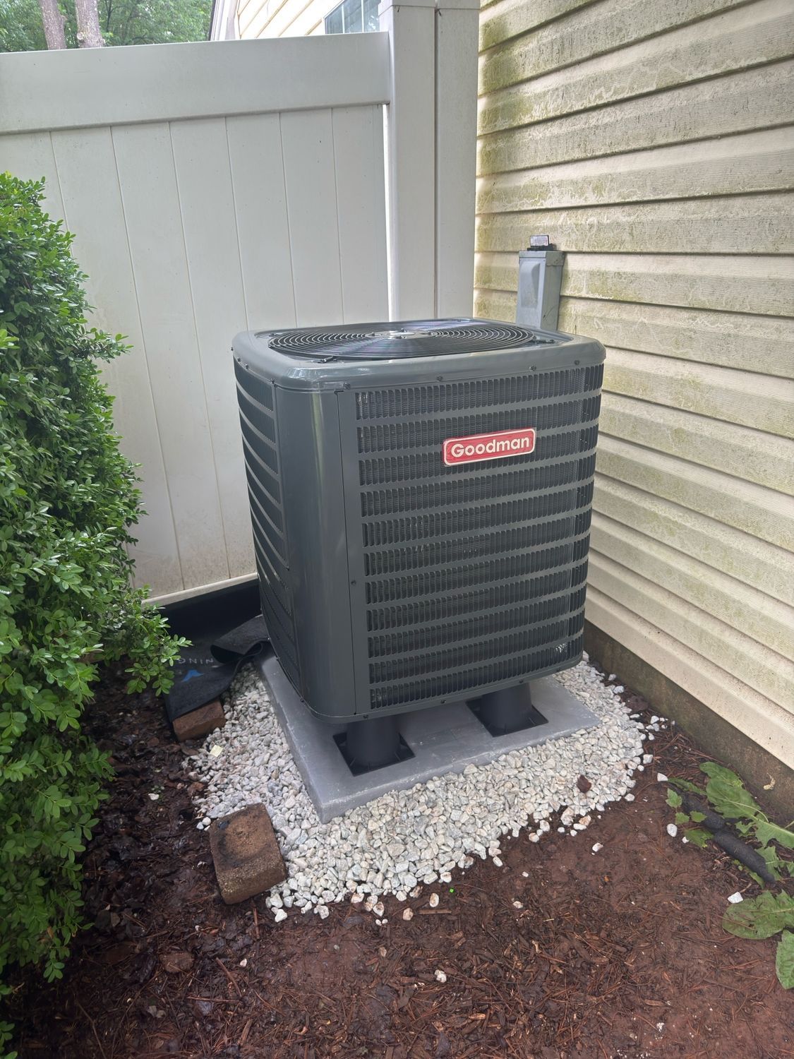 Outdoor HVAC condenser unit on a concrete pad beside a house, with gravel and shrubs nearby.