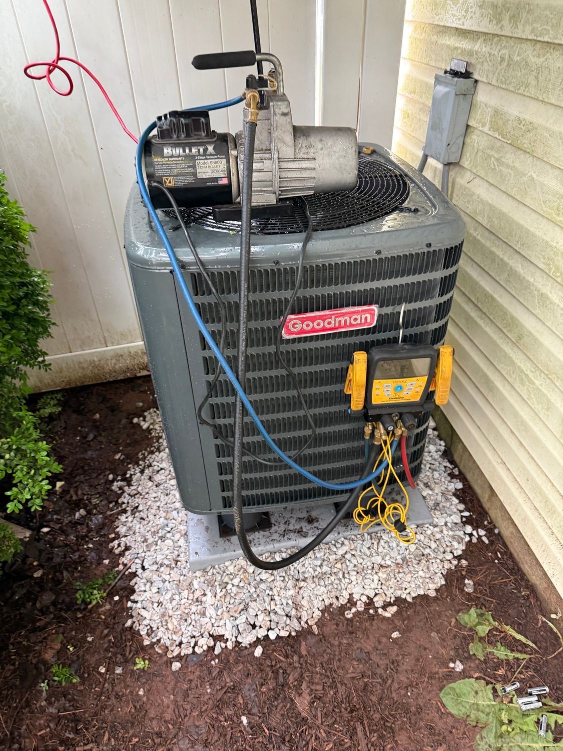 Outdoor air conditioner unit with gauges and hoses attached, set on a gravel bed beside a wall.