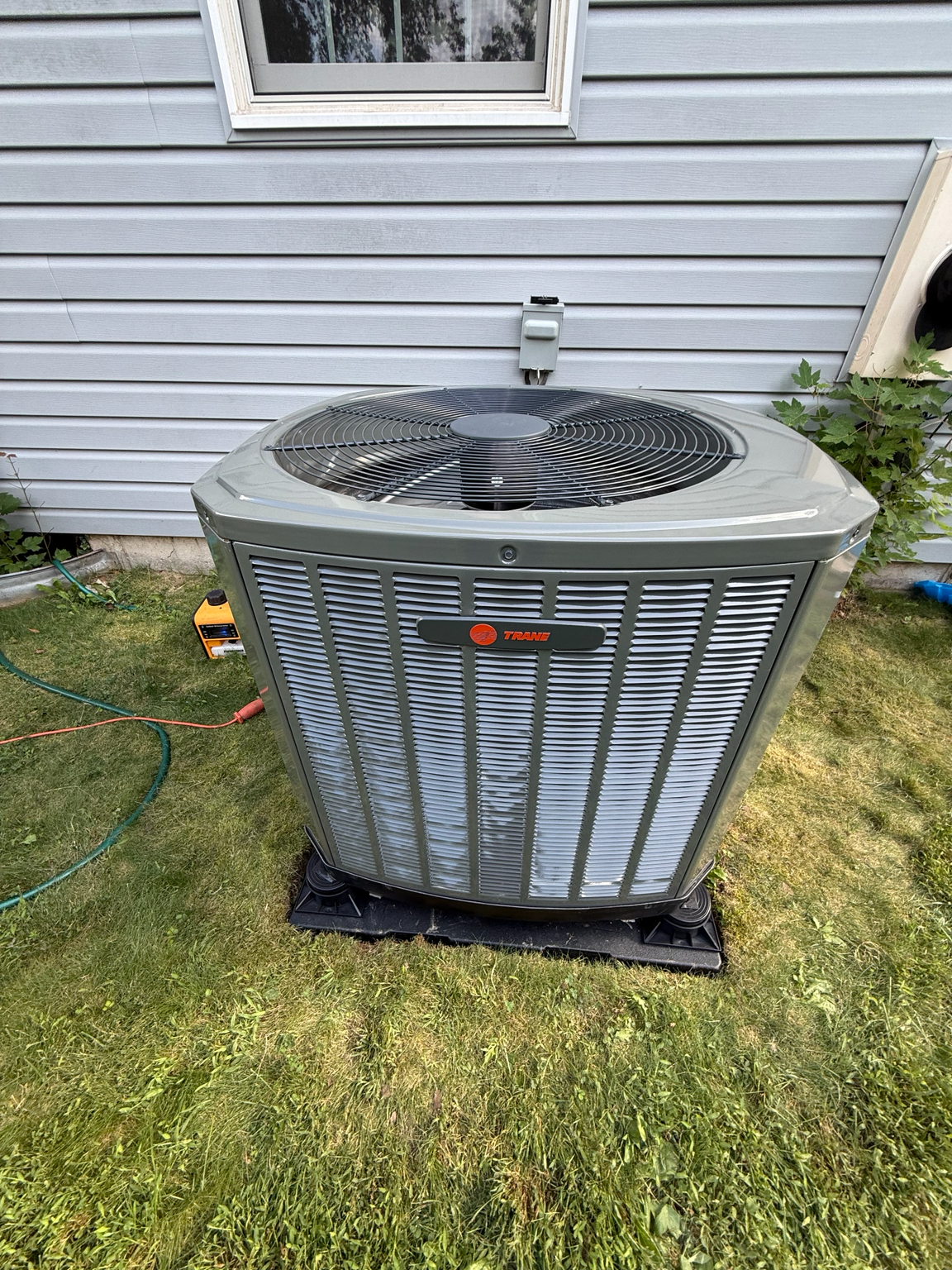 Outdoor air conditioner unit beside a house on a grassy lawn, with a hose and window nearby