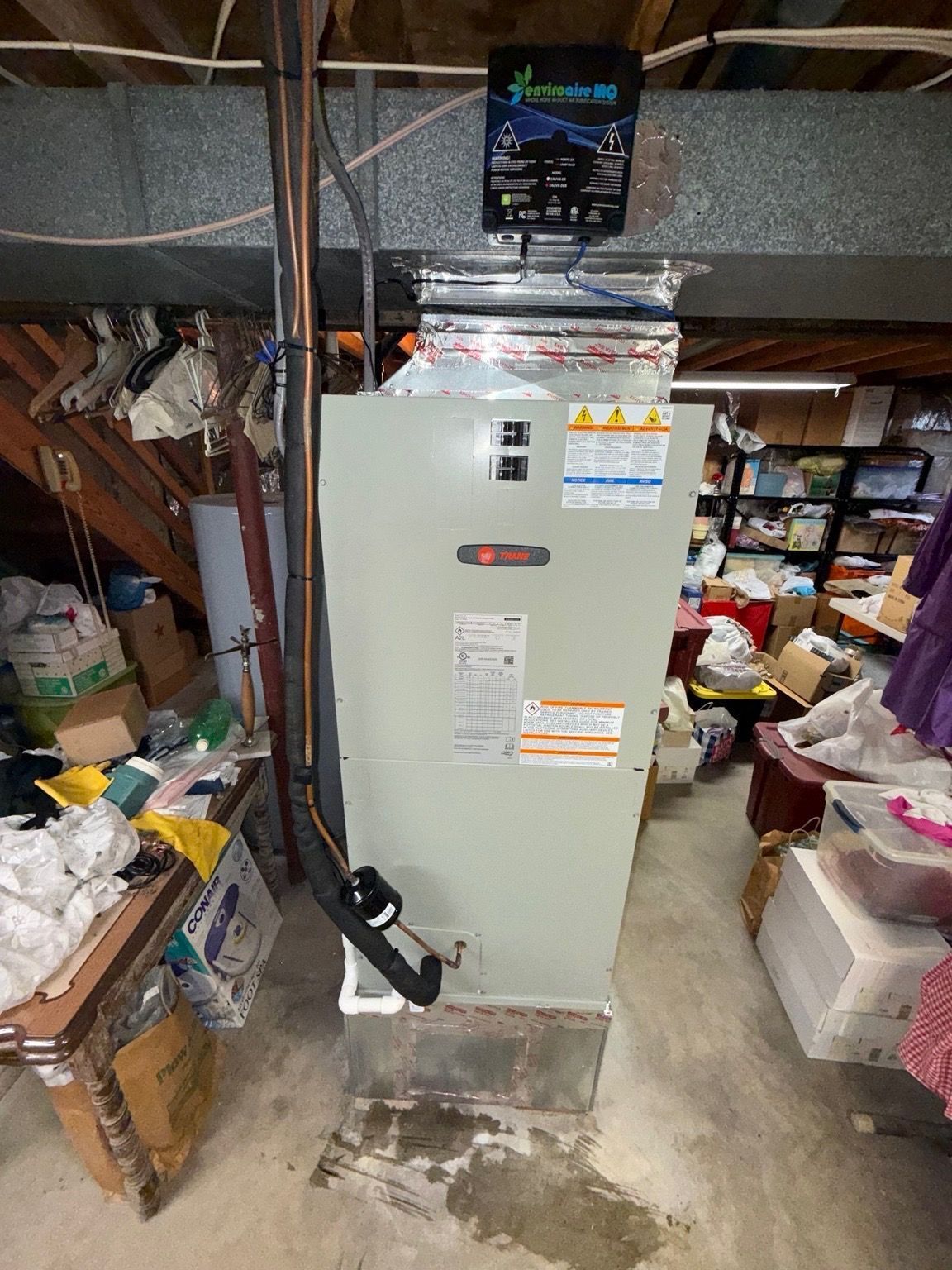 Beige furnace in a cluttered basement utility room with exposed pipes and stored boxes.