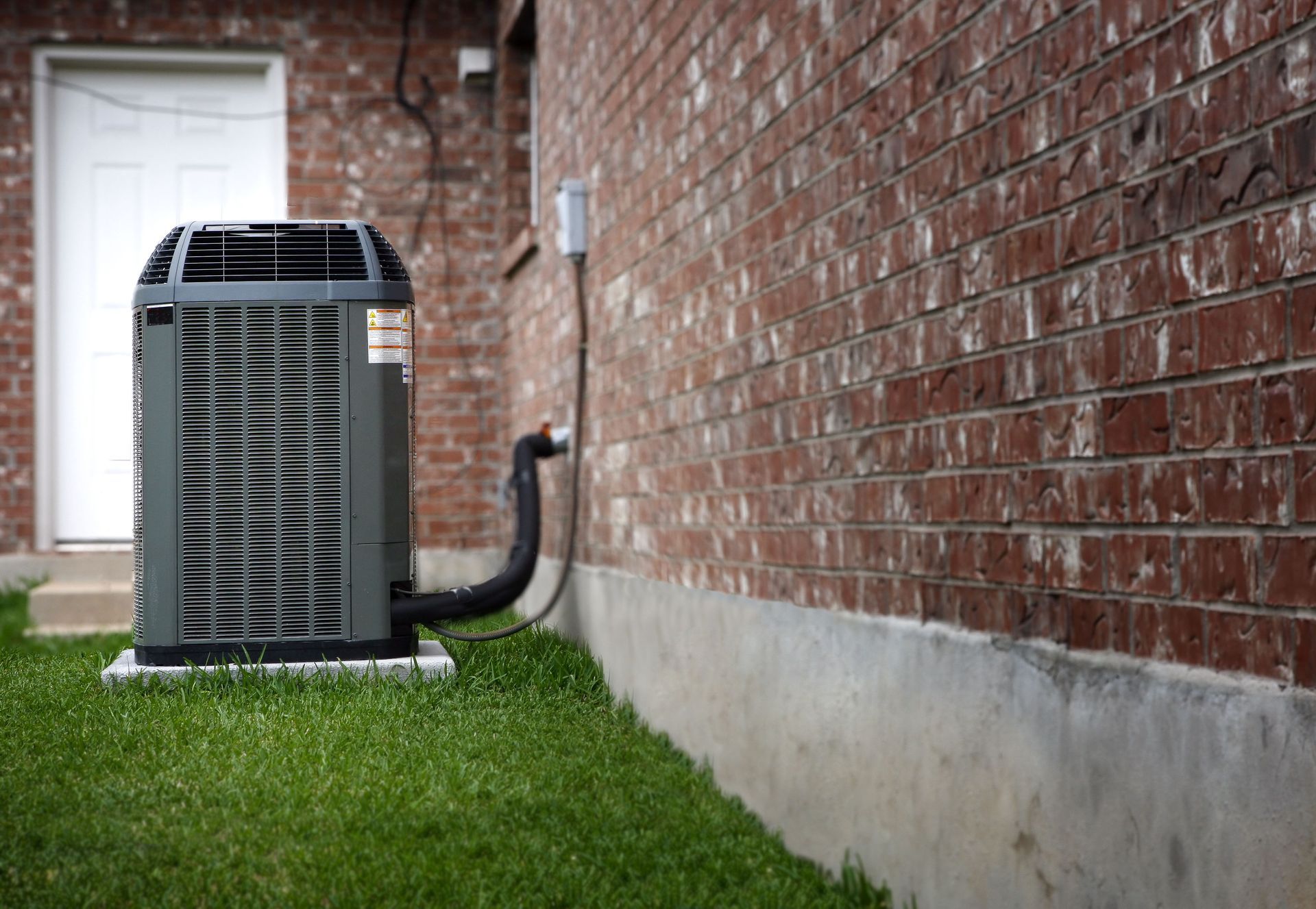 Outdoor air conditioner unit beside a brick house with grass and a window nearby