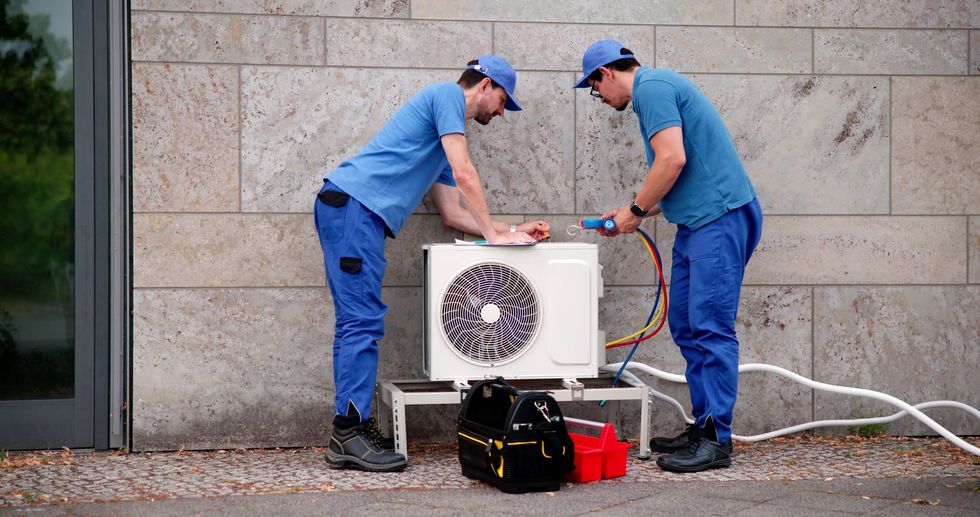 Two technicians in blue uniforms inspecting an outdoor air conditioning unit beside a stone wall.