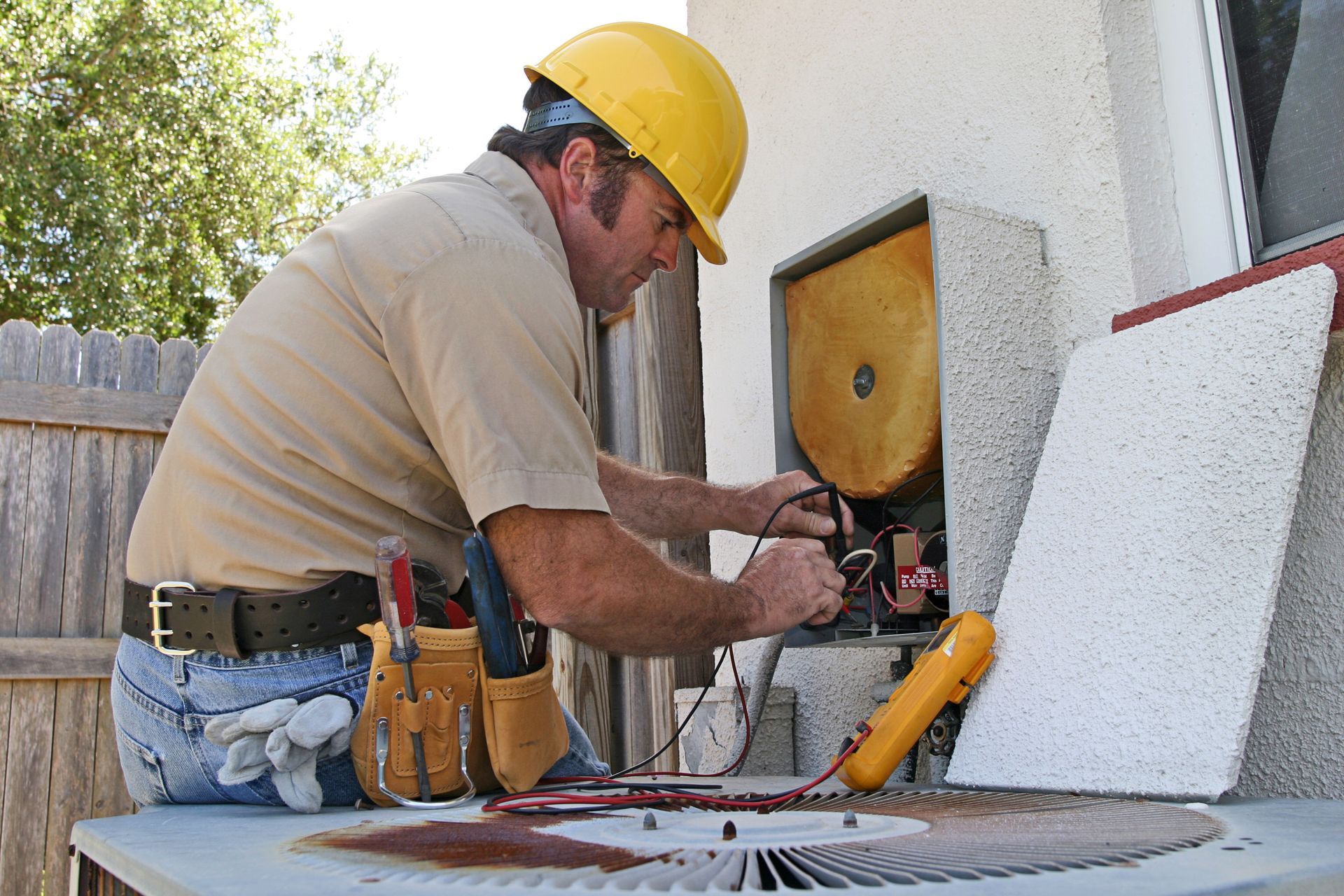 Worker in yellow hard hat cutting material outdoors beside tools and a house wall