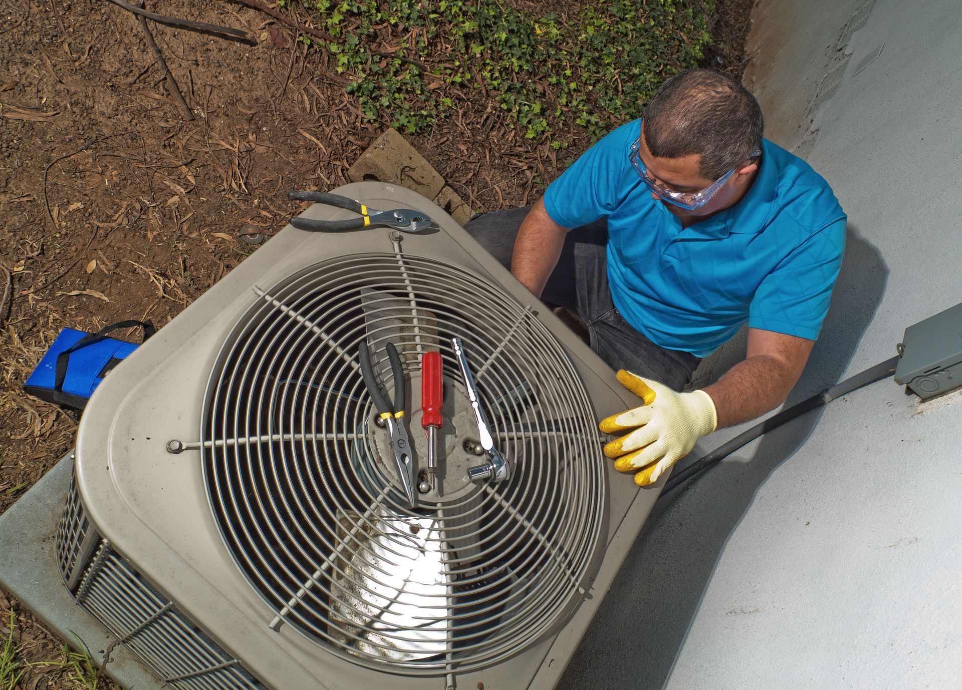Technician in blue shirt inspecting an outdoor HVAC unit with a red tool and yellow gloves