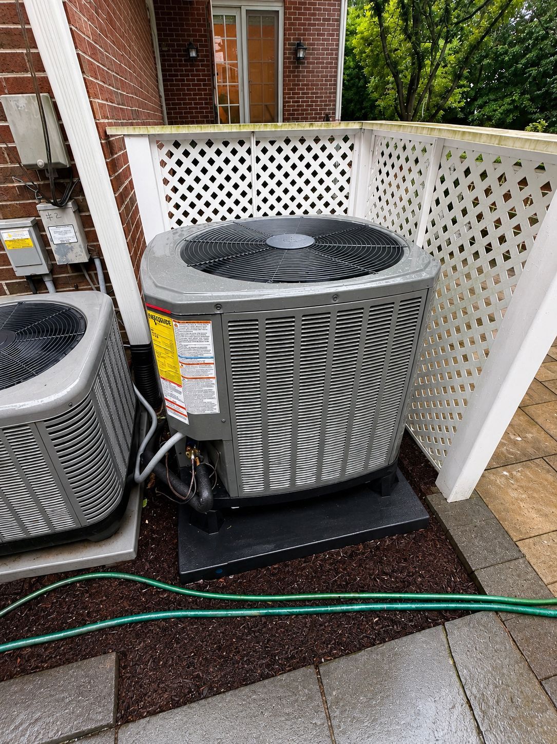 Outdoor air conditioner units beside a brick house, fenced patio, and wet ground with hoses.