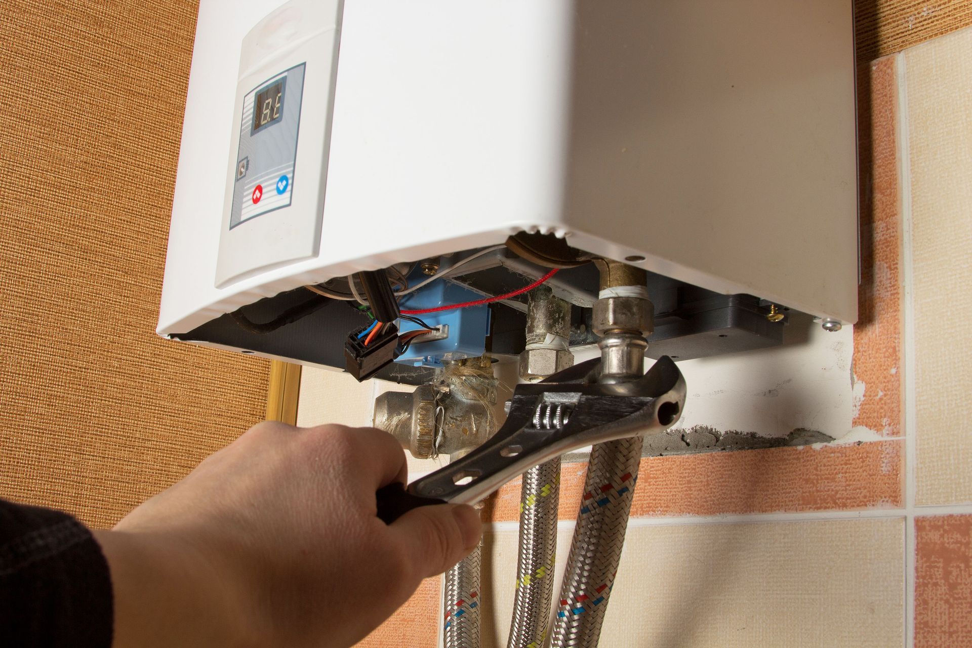 Hand adjusting plumbing under a white wall-mounted water heater with exposed pipes and hoses
