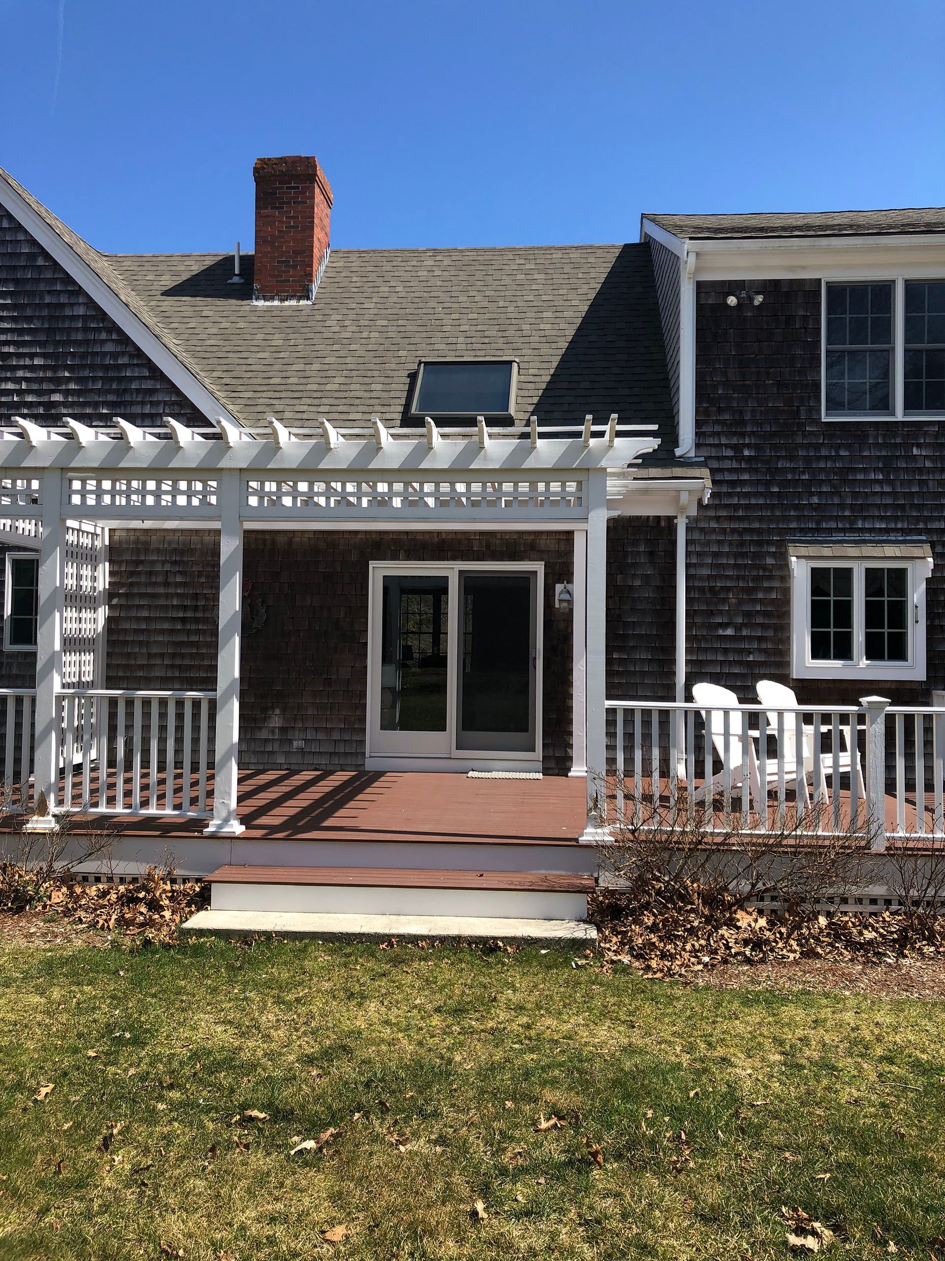 Back of weathered shingle-sided house with a deck and white pergola, a sliding glass door, and a brown roof.