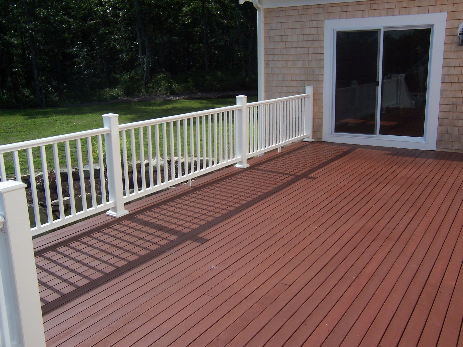 Red deck with white railing next to a building with sliding glass doors, green lawn in background.