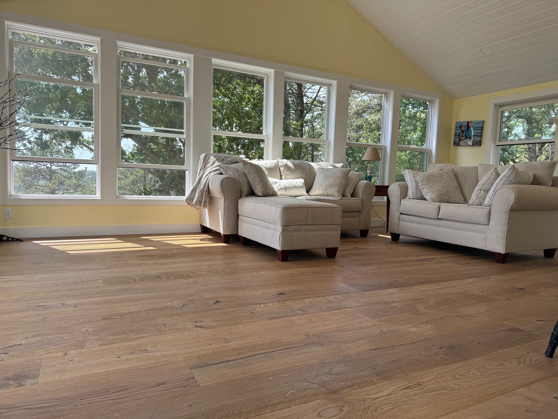 Sunlit living room with light-colored hardwood floor, cream-colored sofas, and large windows overlooking trees.