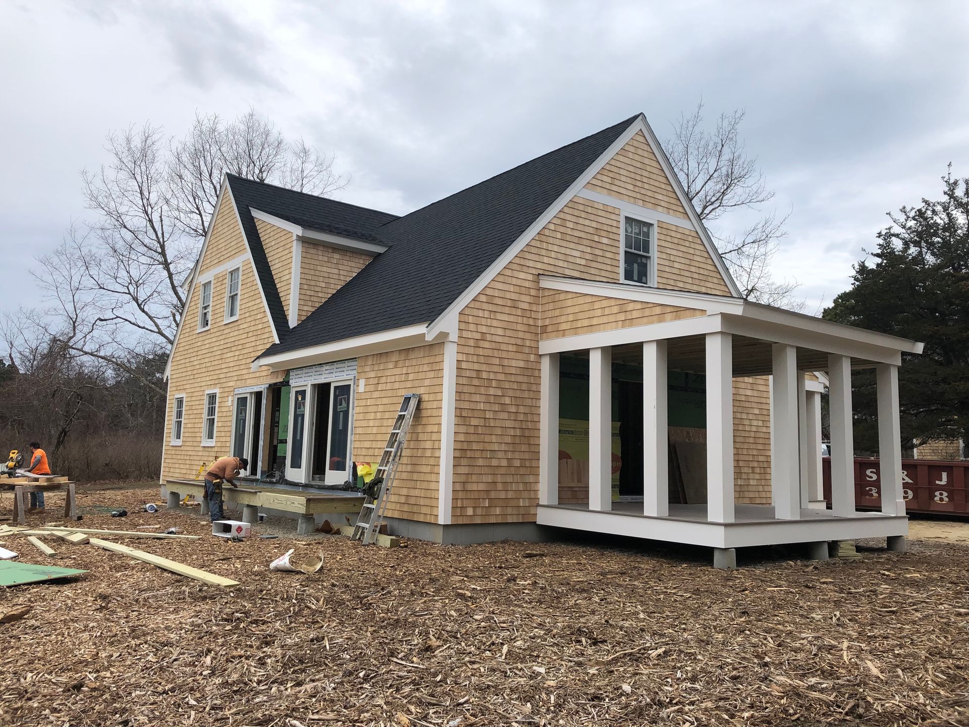 House under construction with cedar siding and a porch. Workers are visible outside.
