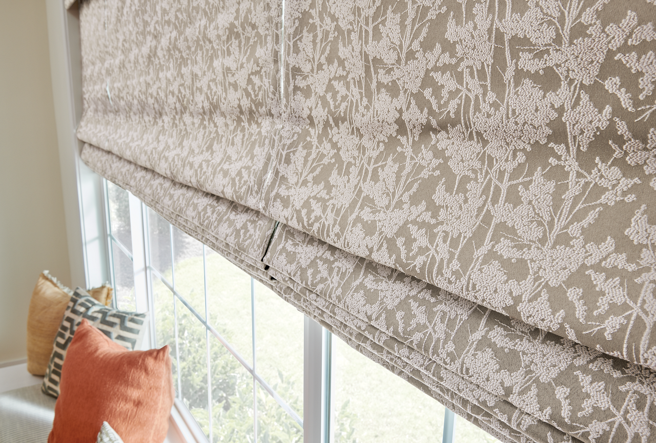 Patterned beige Roman shade over a window with orange pillows below