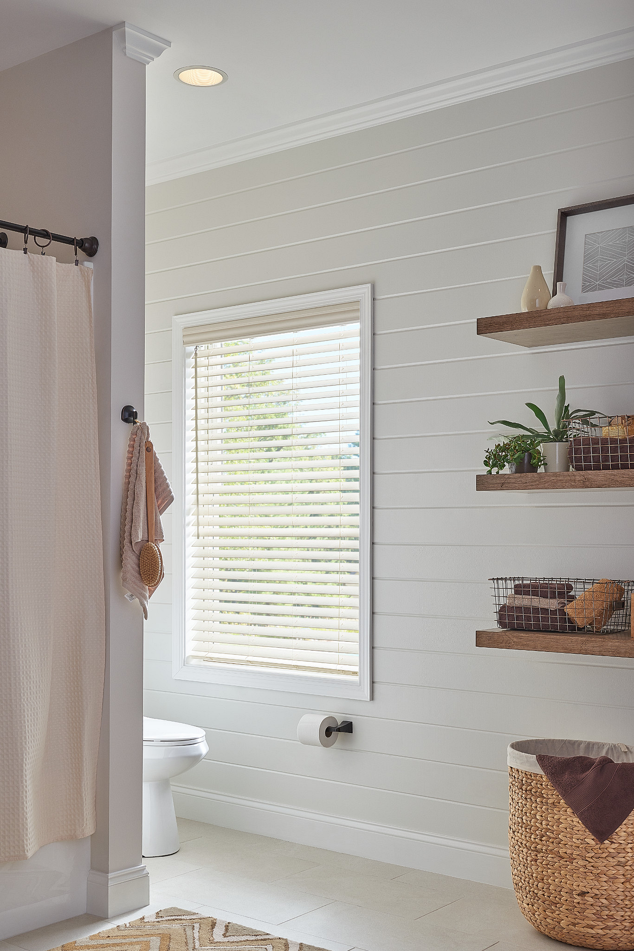 Bright bathroom with white shiplap walls, window blinds, towel hook, shelf decor, and a woven basket.