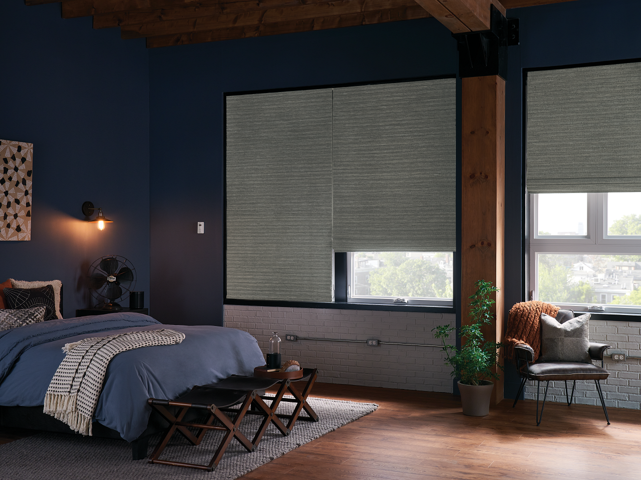 Modern bedroom with navy walls, a bed, woven stools, and large windows with blinds and a warm lamp glow