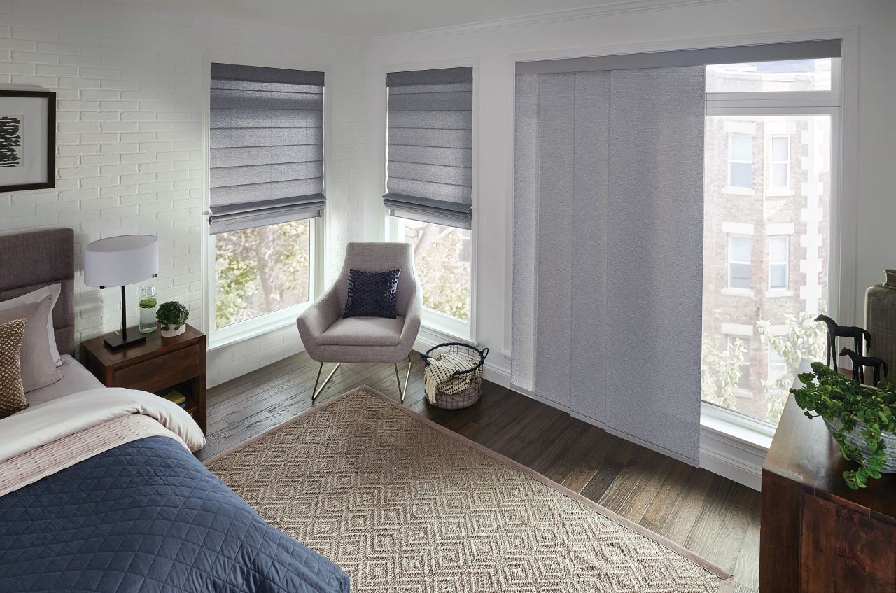 Bright bedroom with large windows, beige rug, armchair, bed, and plants by the window.