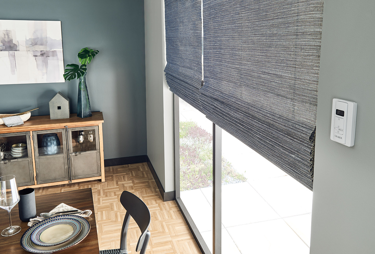 Dining area with a woven table, gray walls, and a large window covered by a textured roller shade.