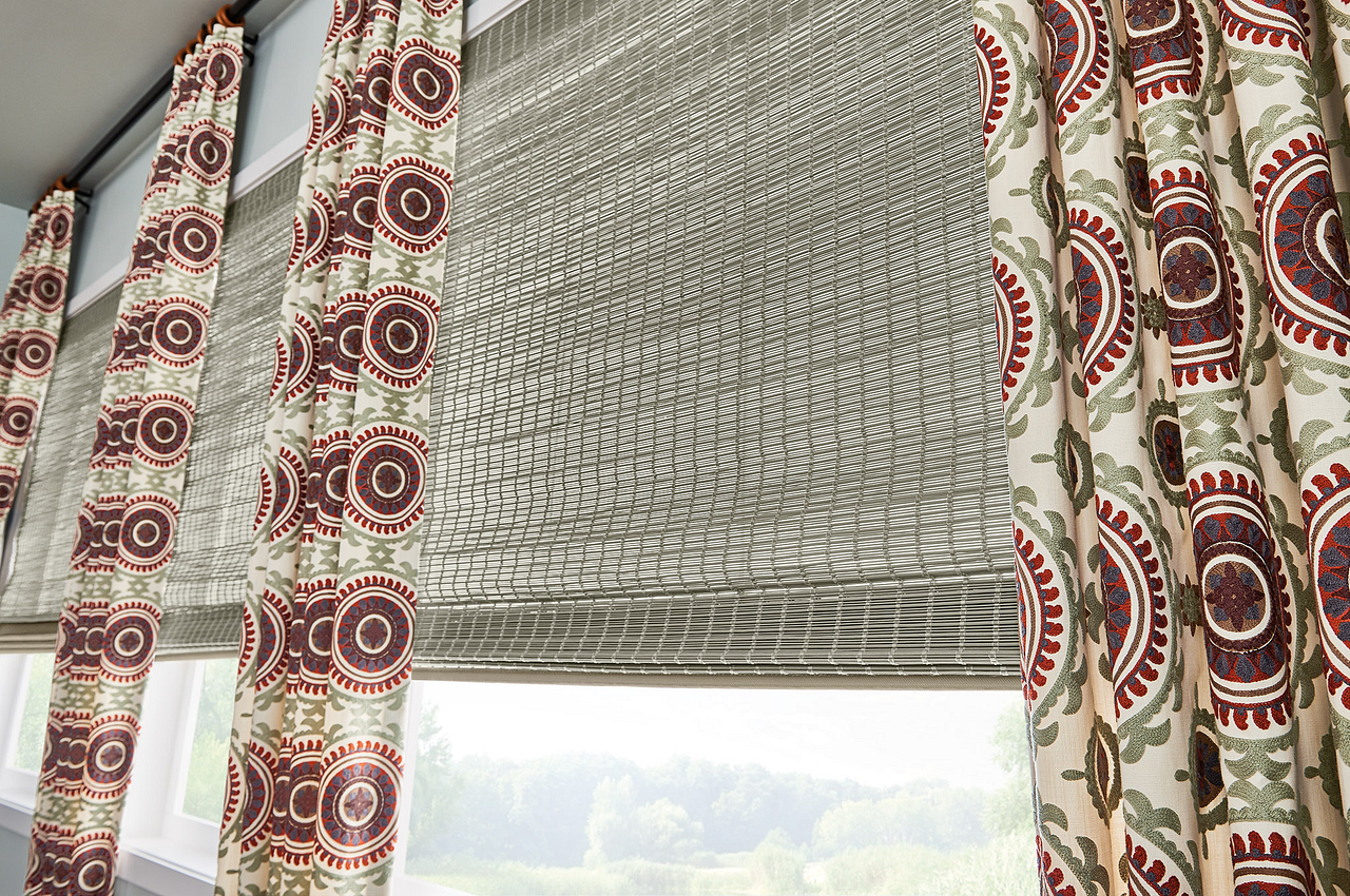 Patterned curtains frame a window with a woven bamboo shade partially raised.