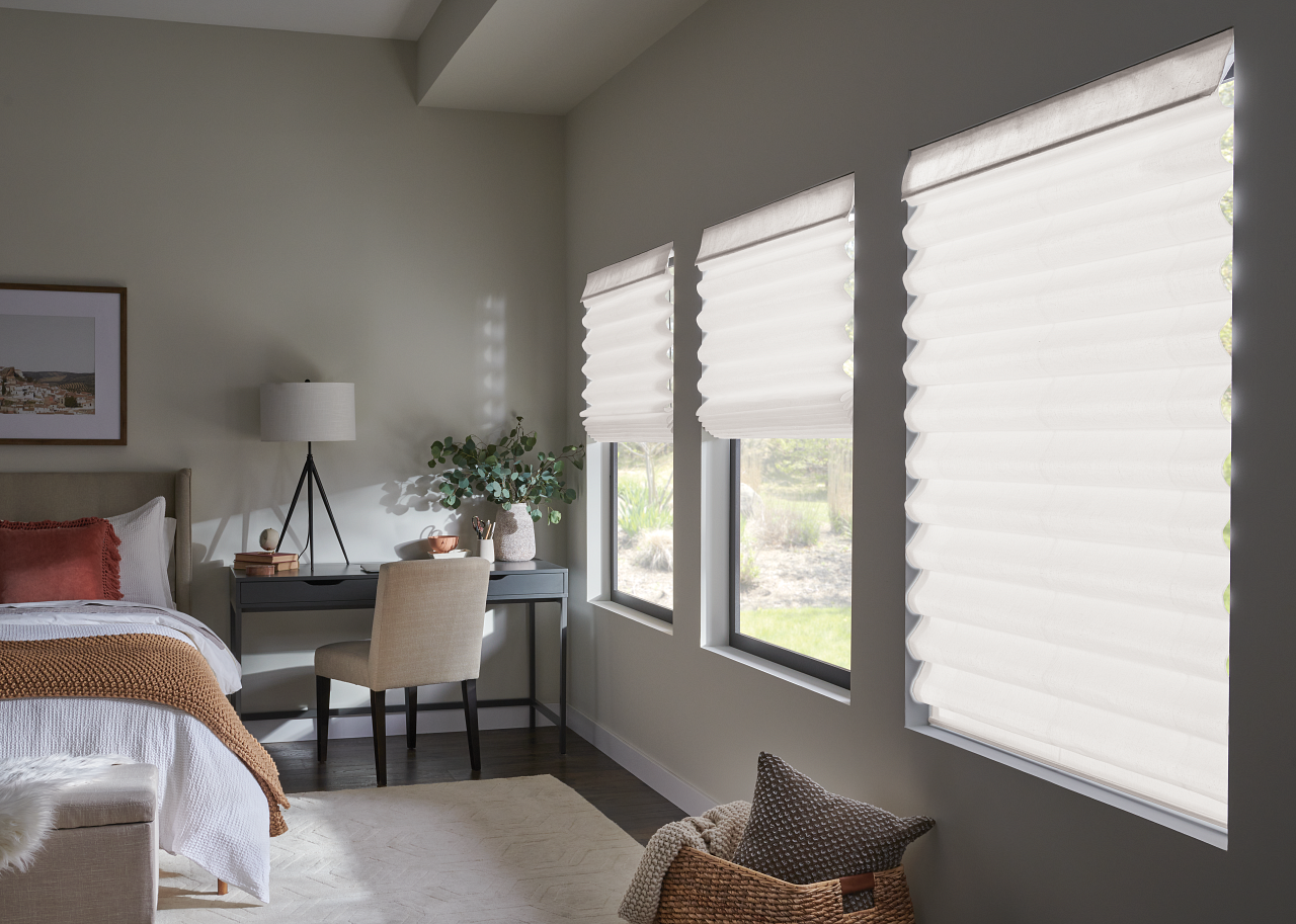 Bright bedroom with a bed, desk, chair, and large windows with white blinds.