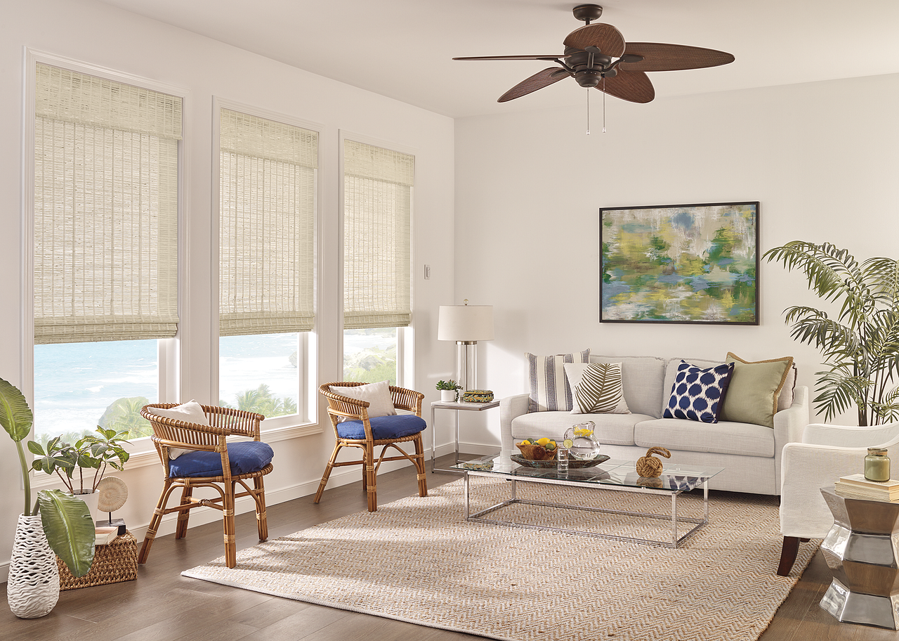 Bright living room with beige sofa, woven chairs, large windows, rug, plants, and ceiling fan