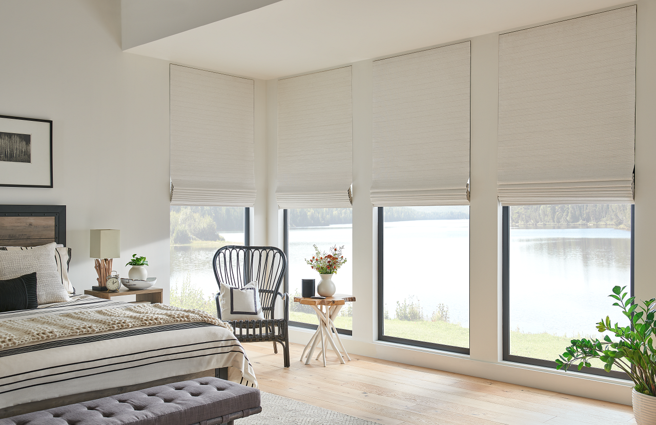 Bright bedroom with large windows, white blinds, patterned bed, and a small seating area overlooking water