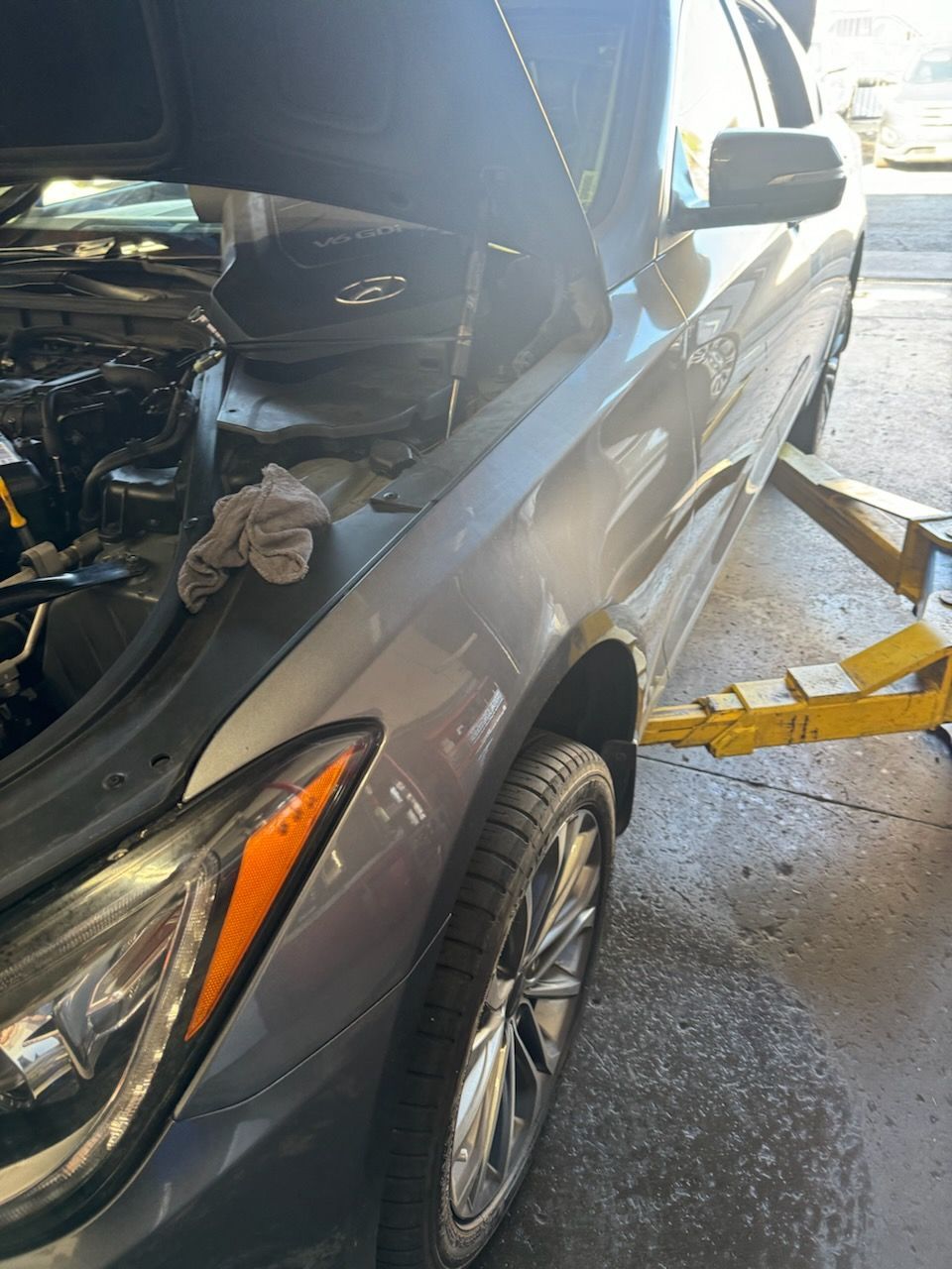 Gray car being worked on in a shop with hood open, propped on a lift, and wheel visible.