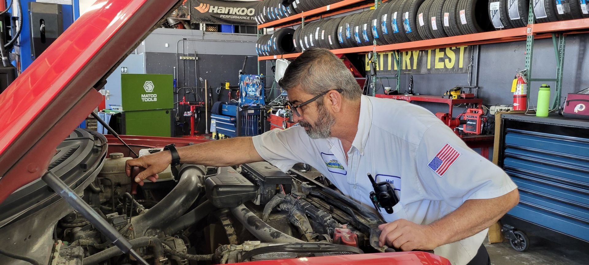 A mechanic in a white shirt with an American flag patch checks a car engine in a garage.