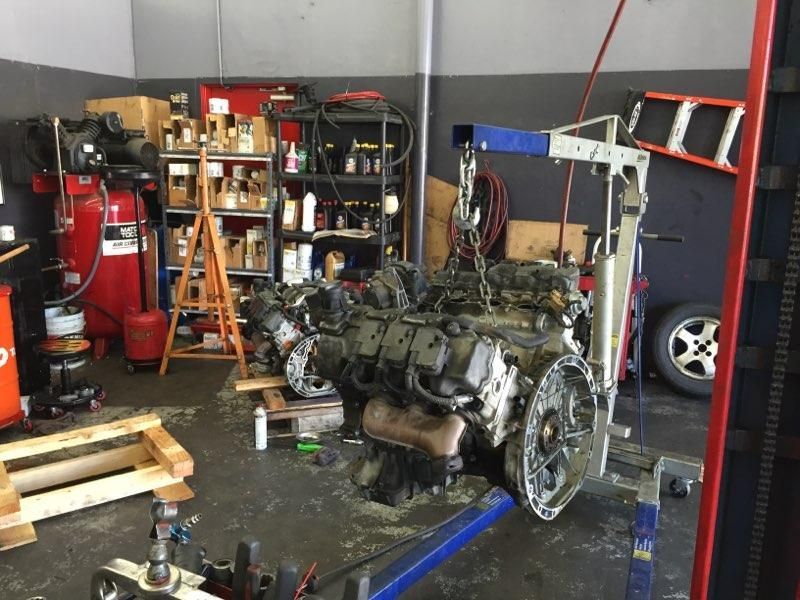 Engine being hoisted in a cluttered auto repair shop, with tools, shelves of fluids, and a red air compressor.