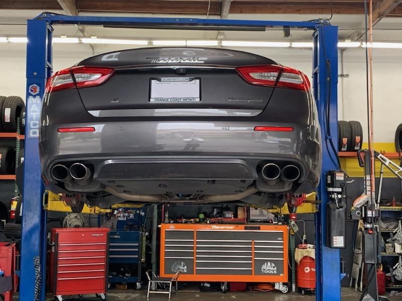 Gray Maserati sedan on a car lift in a shop, rear view showing exhaust pipes and toolbox.