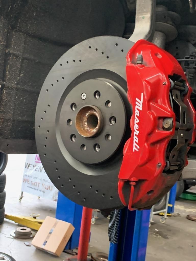 Red Maserati brake caliper and drilled rotor on a car lift.