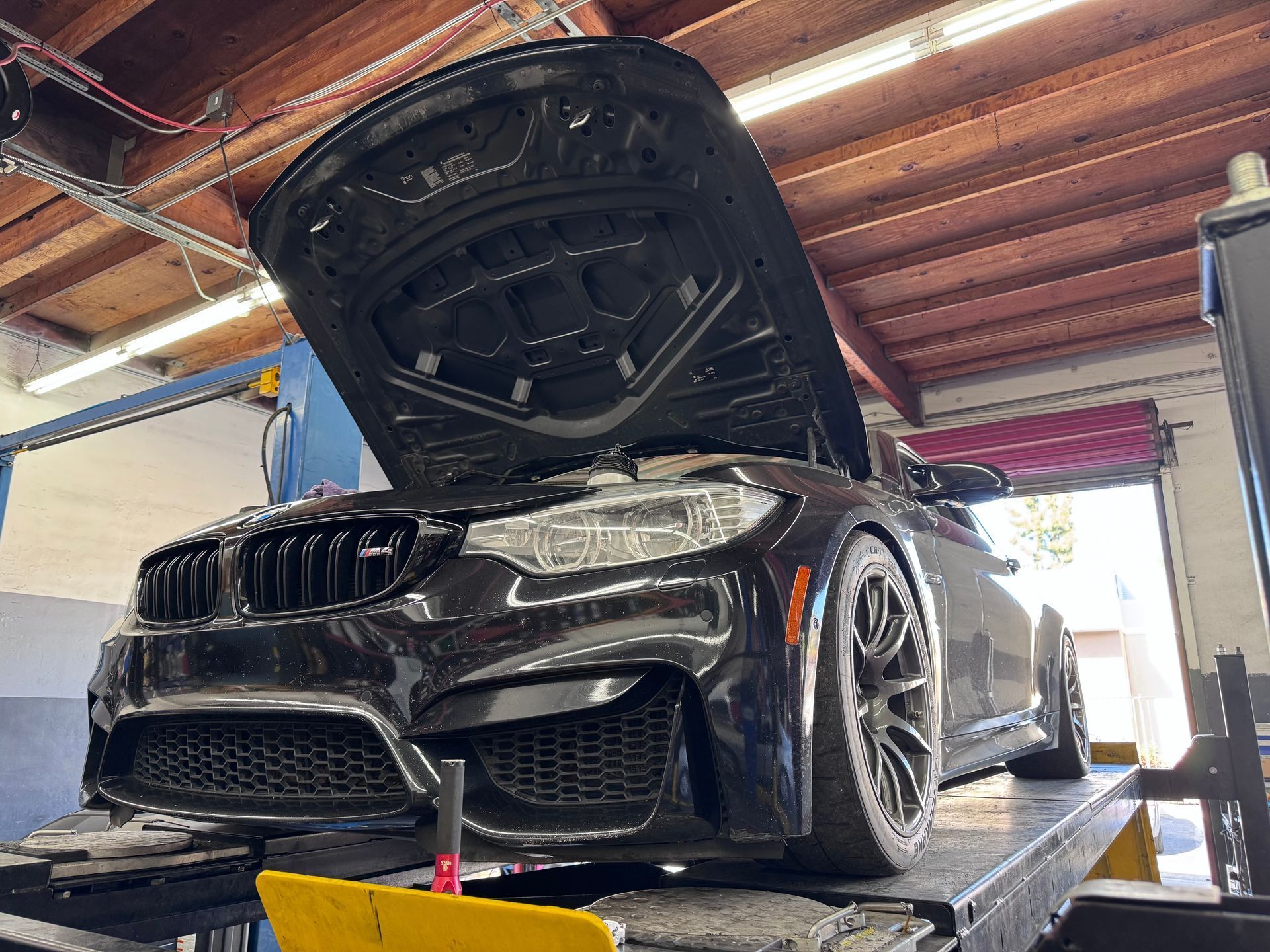 Black BMW sports car on a lift with hood open in a garage, showing the front end and engine bay.