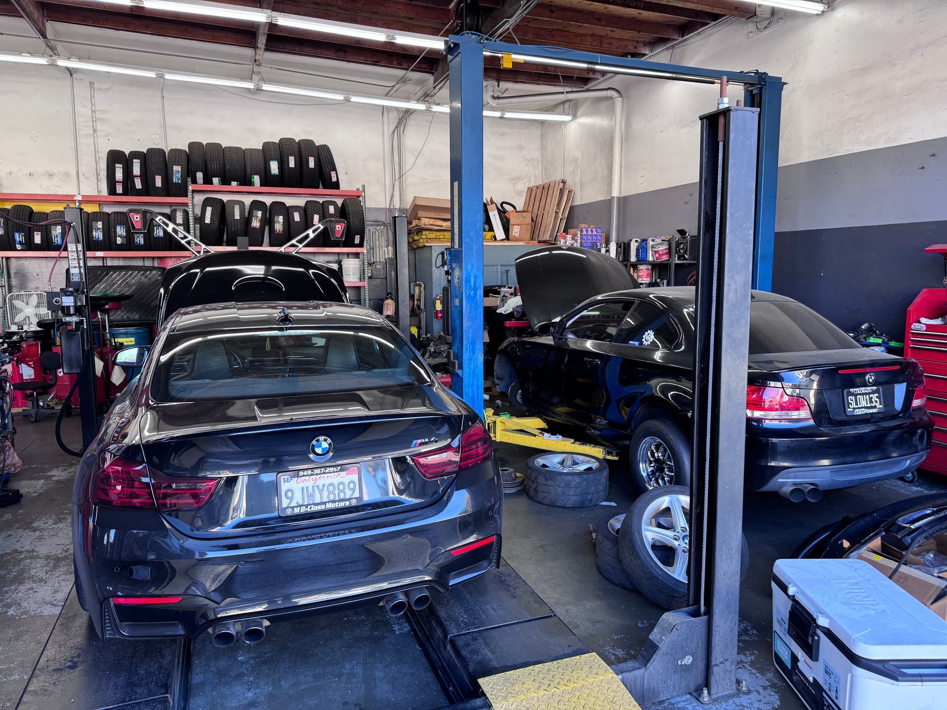 Two black sedans in an auto repair shop with one car raised on a lift and shelves of tires in the background.