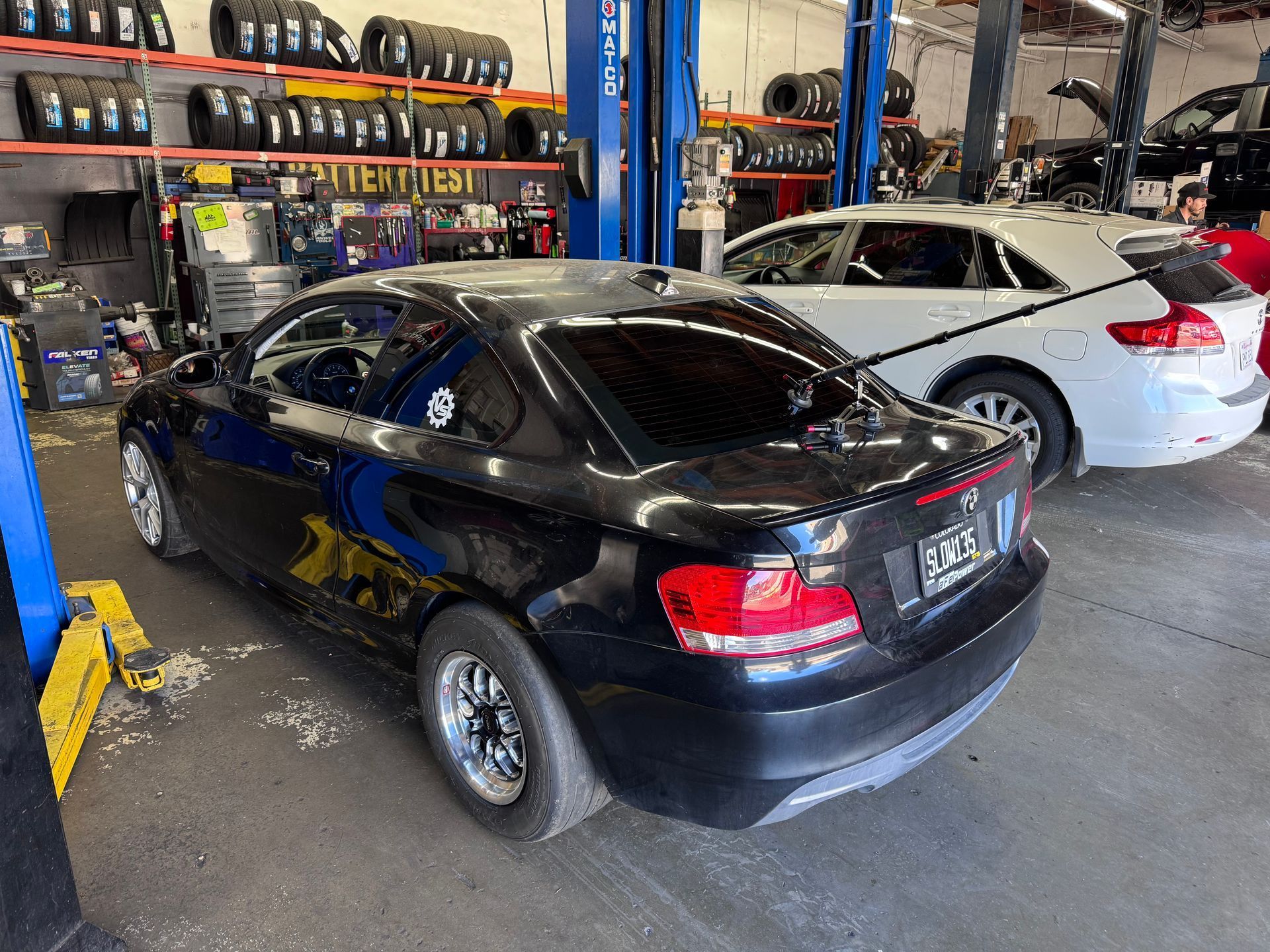 Black sedan and white coupe parked inside an auto repair shop with tires and tools in the background.