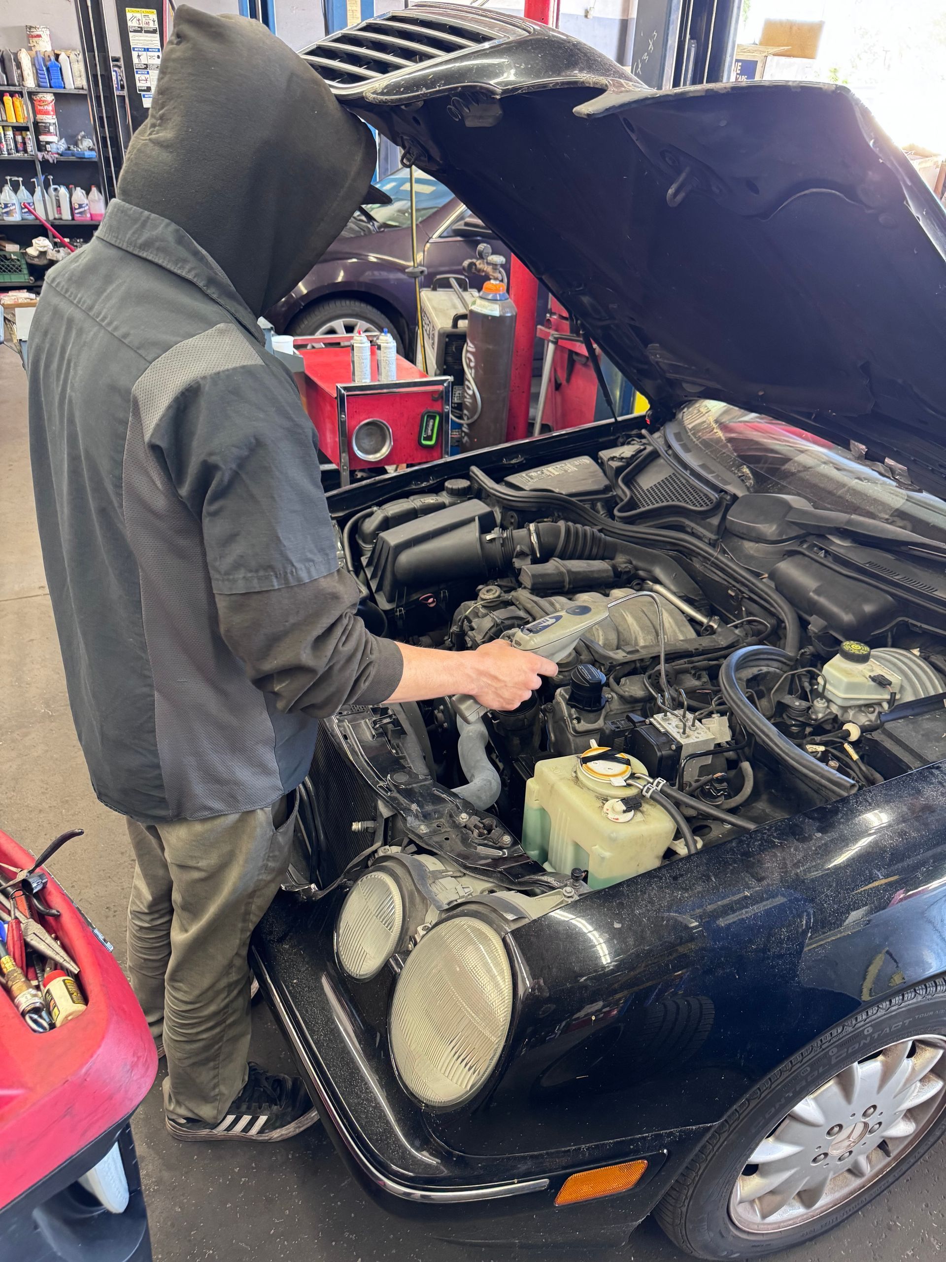 Person works on a black car with hood open in a garage, inspecting the engine.