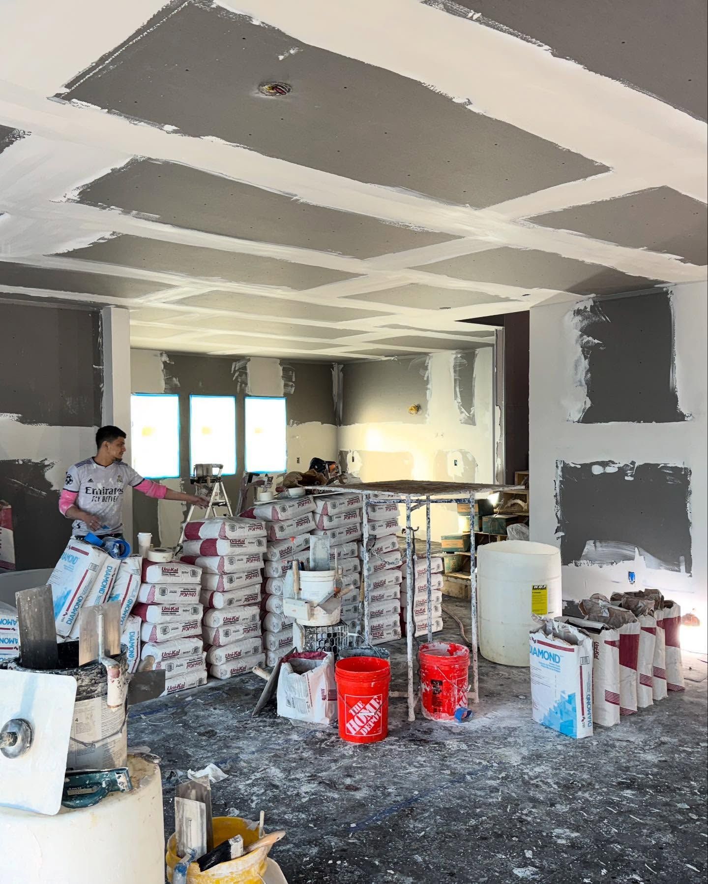 A construction site with a worker surrounded by drywall bags, tools, and partially finished walls and ceiling.