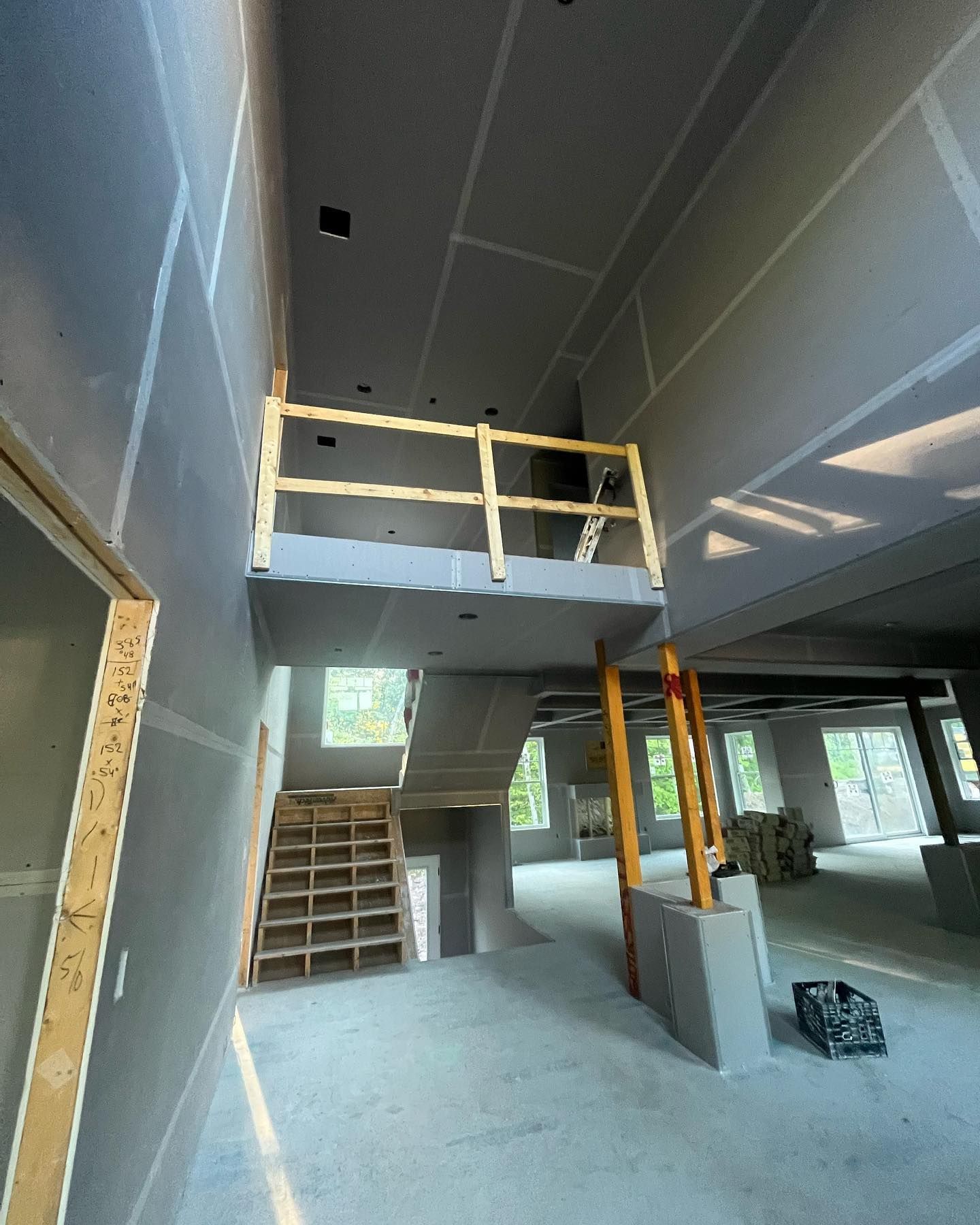 Interior under construction: staircase, mezzanine with railing, drywall, concrete, and lumber.