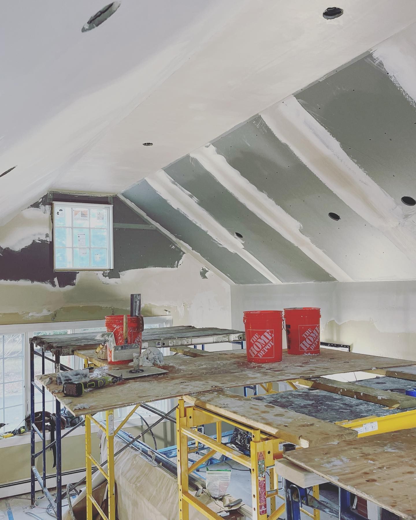 Interior construction: drywall installed on angled ceiling, scaffolding in foreground, buckets of supplies.