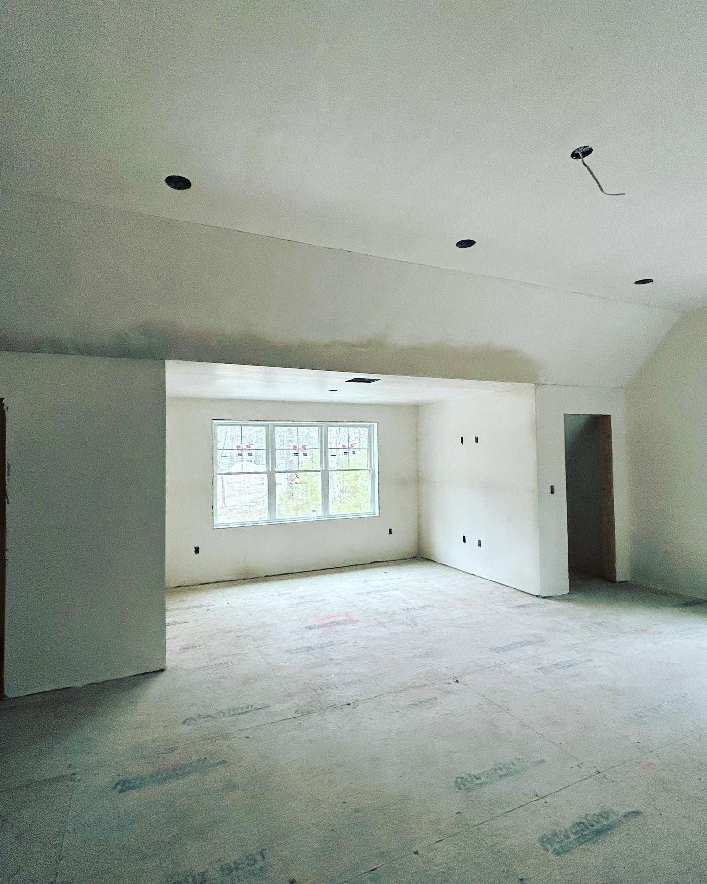 Interior of a room under construction with unfinished walls, ceiling, and concrete floor; a window in the background.