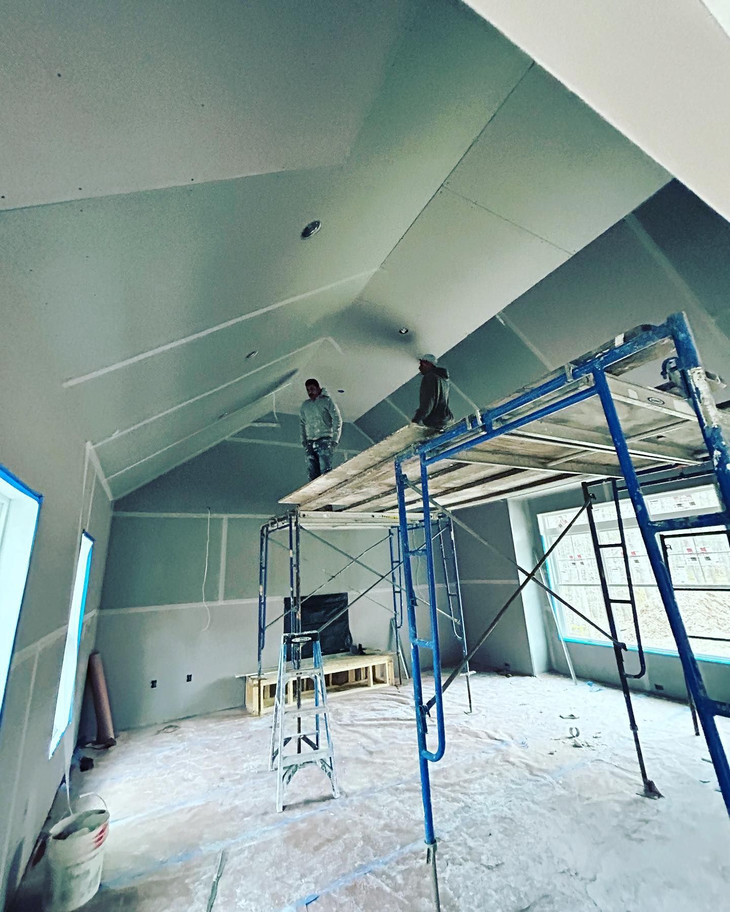 Two workers on scaffolding installing drywall on a sloped ceiling in an unfinished room.