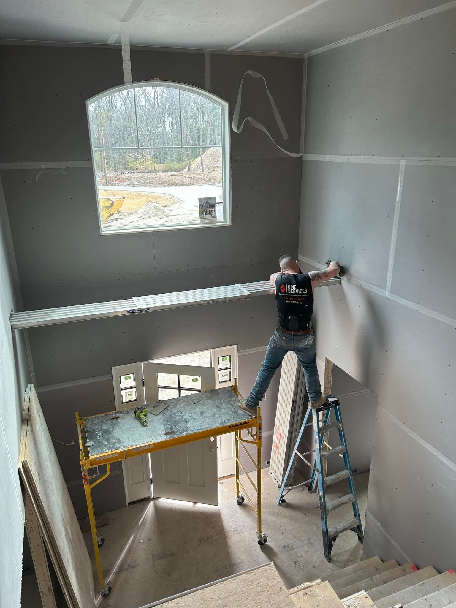 A construction worker on a ladder is working on drywall. The inside of a building under construction with a window in the background.
